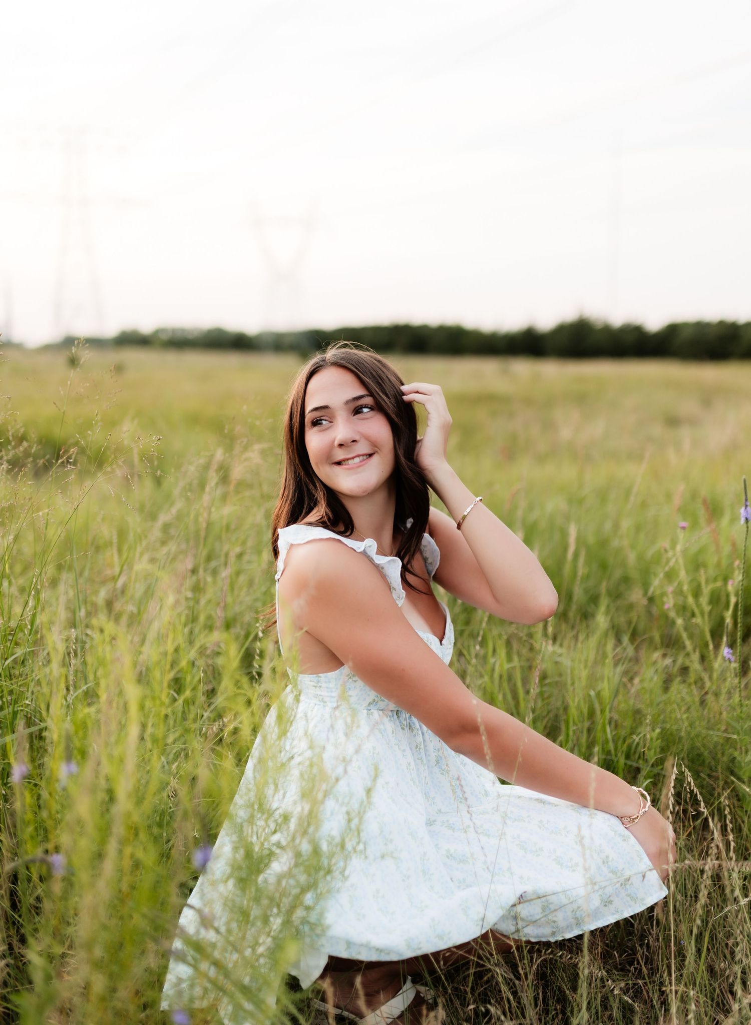 Young woman in white dress sitting in tall wildflower meadow, hand in hair, smiling softly.