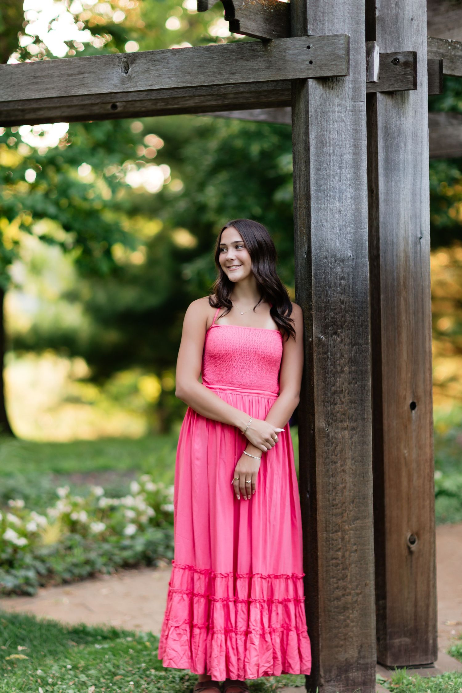 Young woman in bright pink strapless maxi dress leaning against a wooden pergola post in a garden.