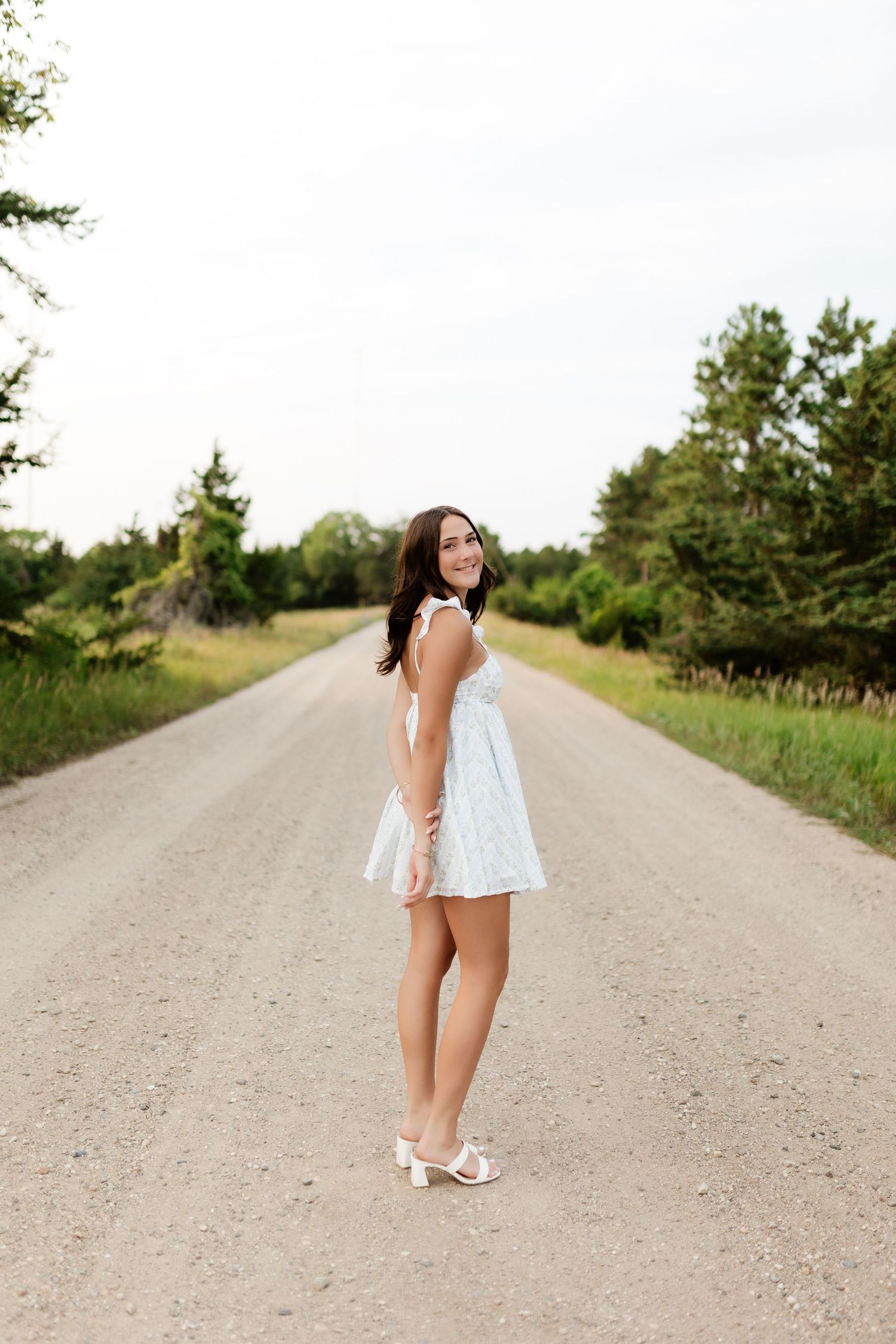 Young woman in white mini dress posing on rural gravel road lined with trees.