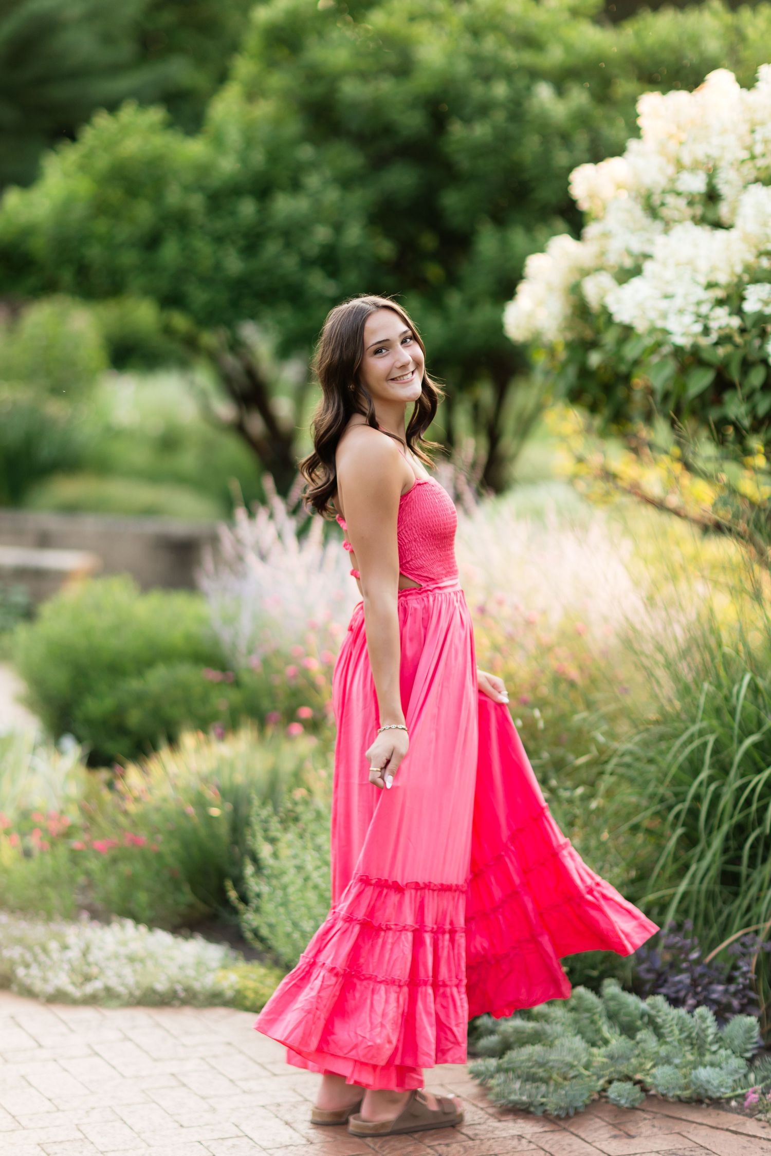 Young woman in coral maxi dress twirling in a lush garden with blooming flowers and greenery.