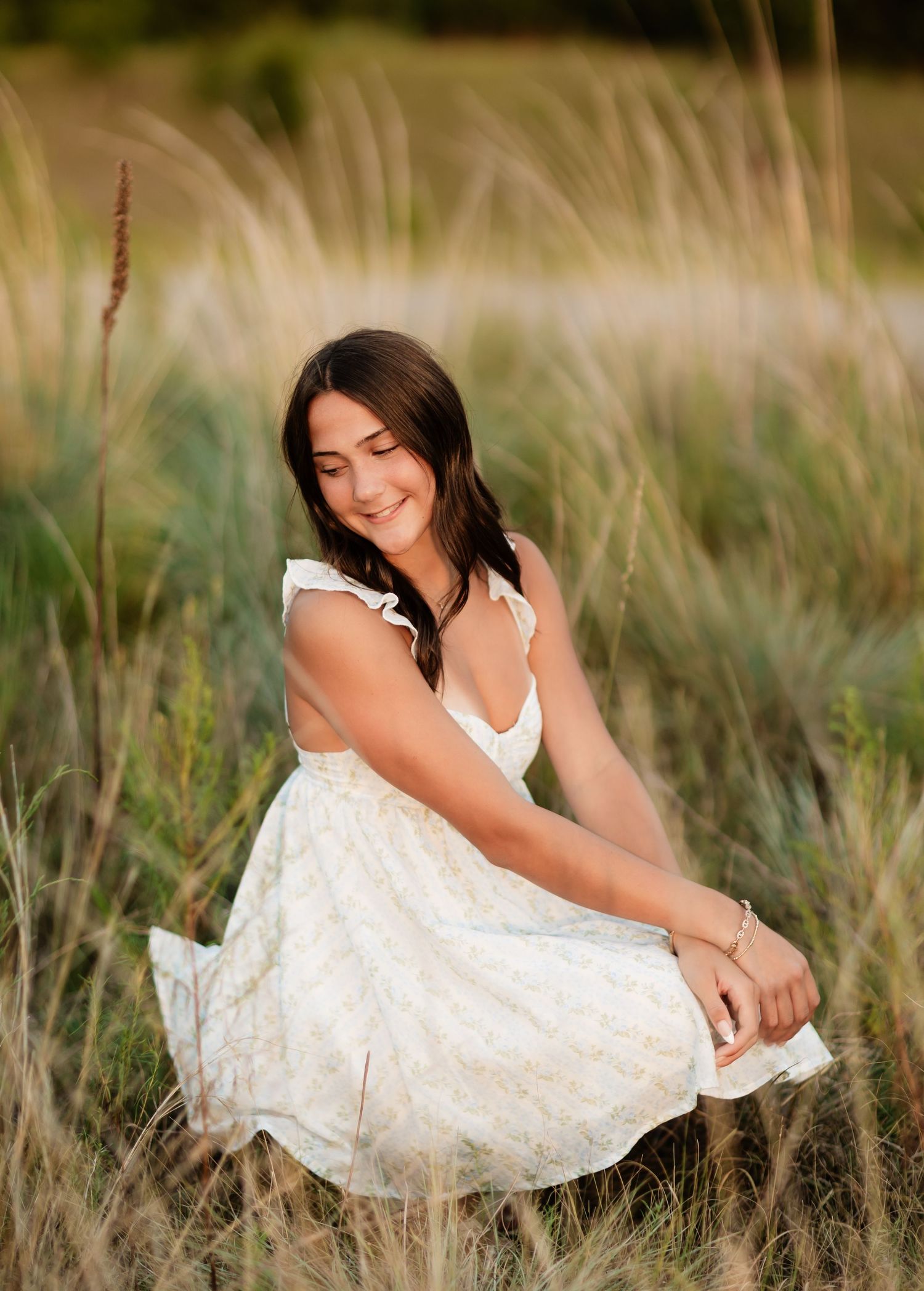 Young woman in white floral dress sitting peacefully among tall golden grasses in soft light.