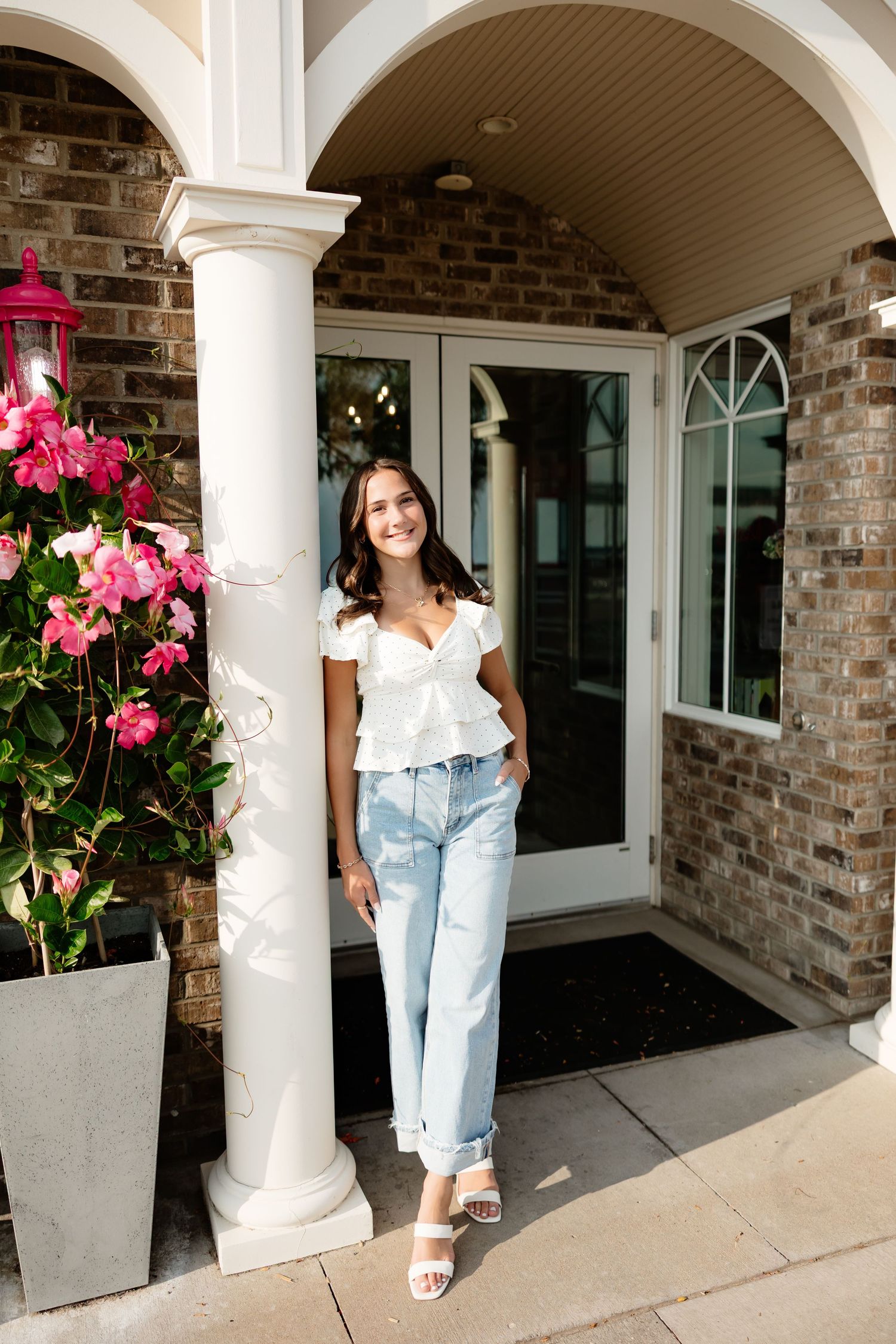 Smiling woman in white top and jeans standing by white column at brick building entrance with pink flowers.