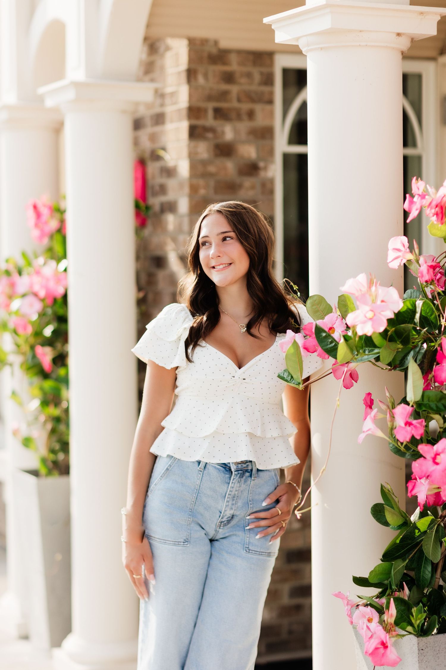 Smiling young woman in white ruffle top and jeans standing between white columns with pink flowers.