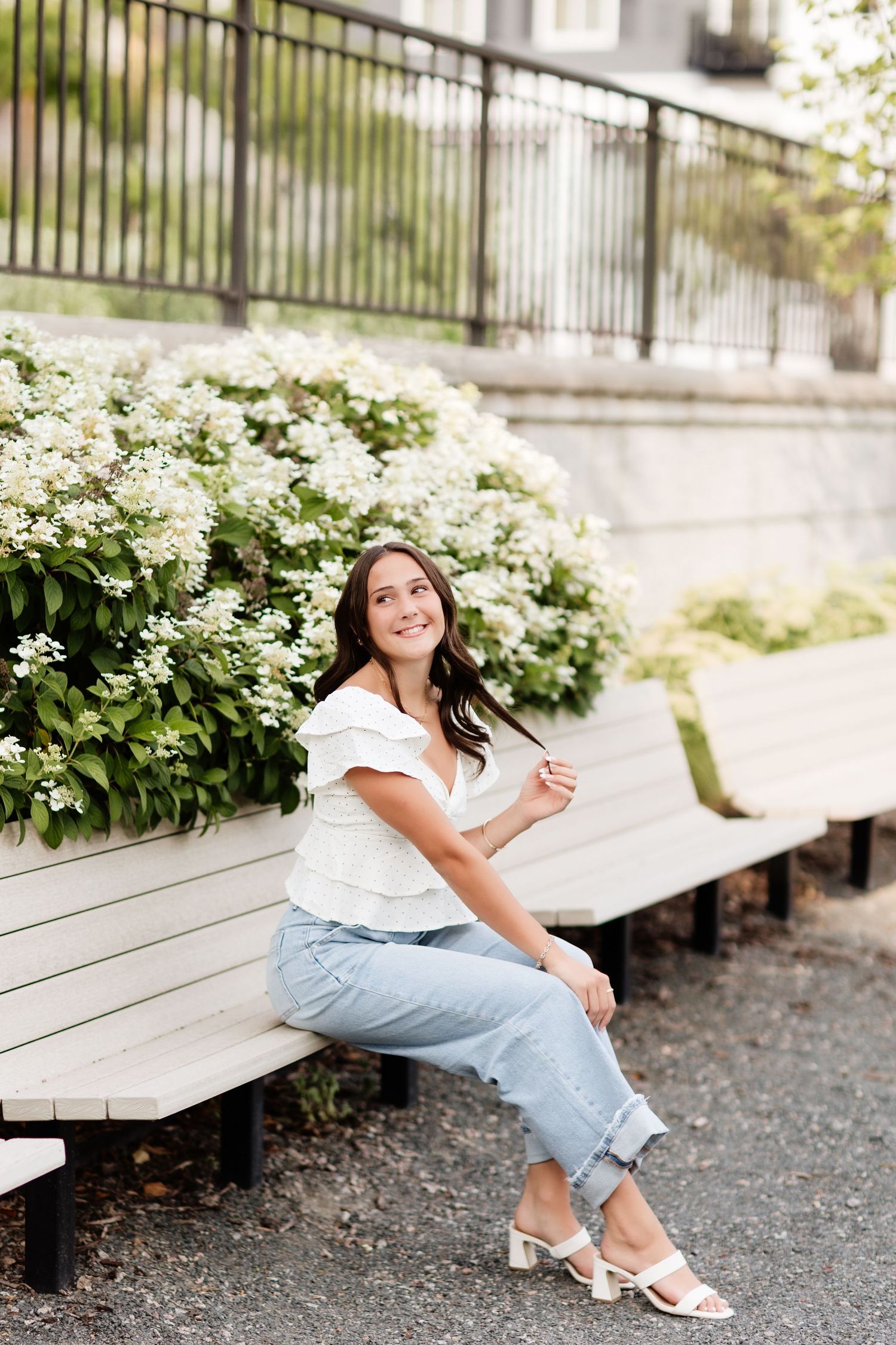 Young woman in white ruffle top and light jeans sits on bench near white flowering bushes, smiling outdoors.
