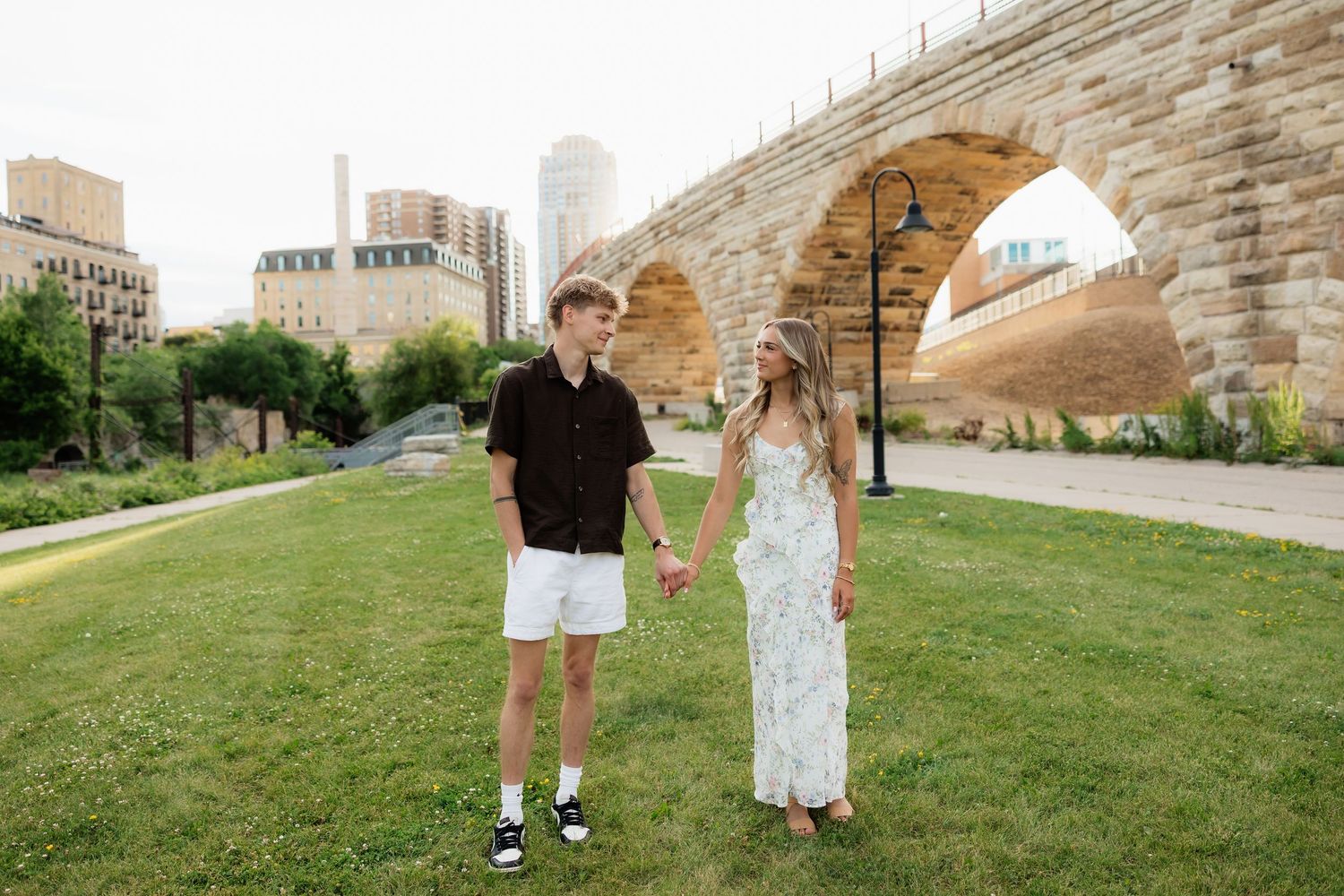 Couple holding hands on green lawn near Stone Arch Bridge in Minneapolis with city skyline in background.