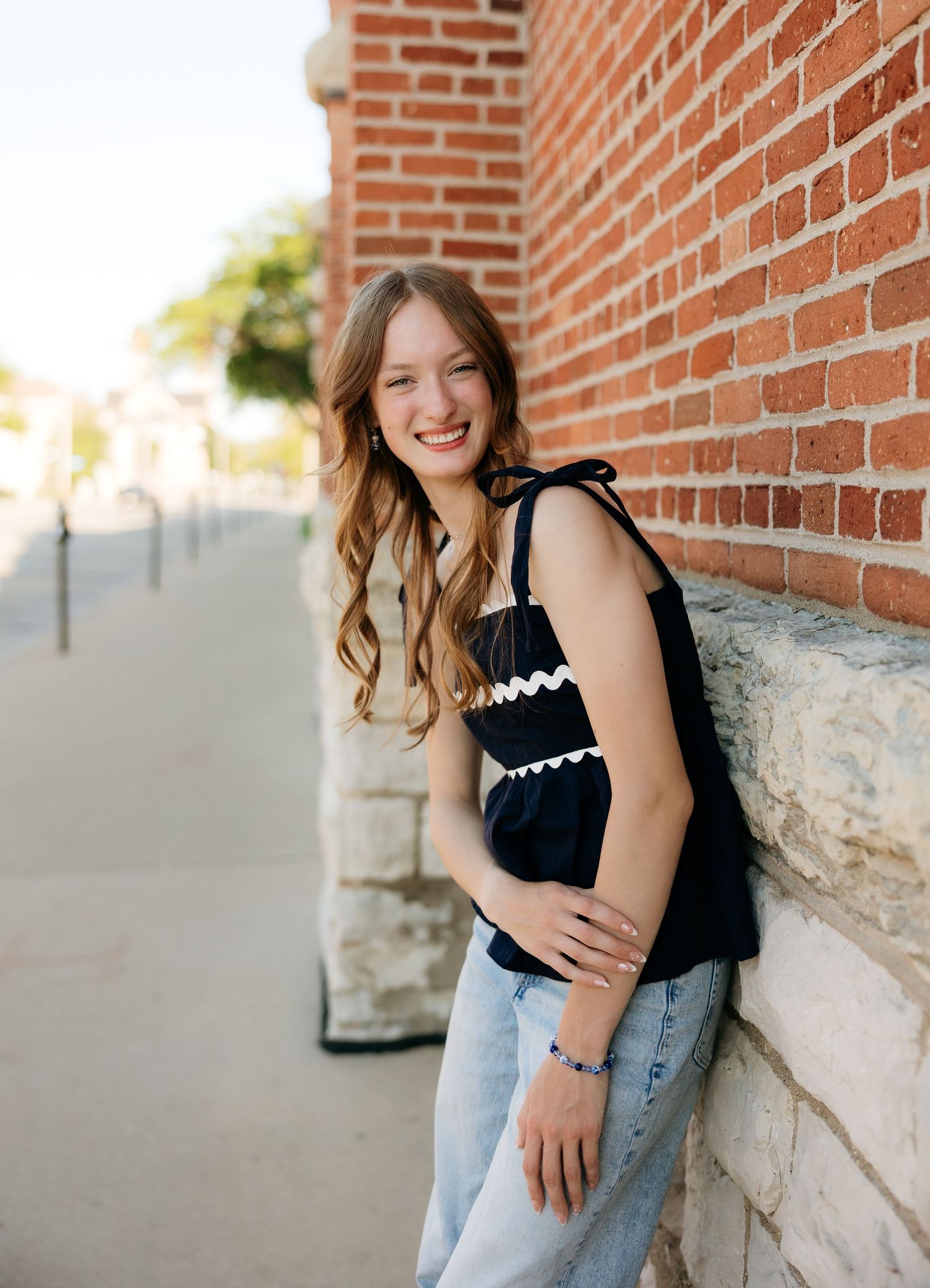 Young woman in black top and jeans smiling against a brick wall outdoors.