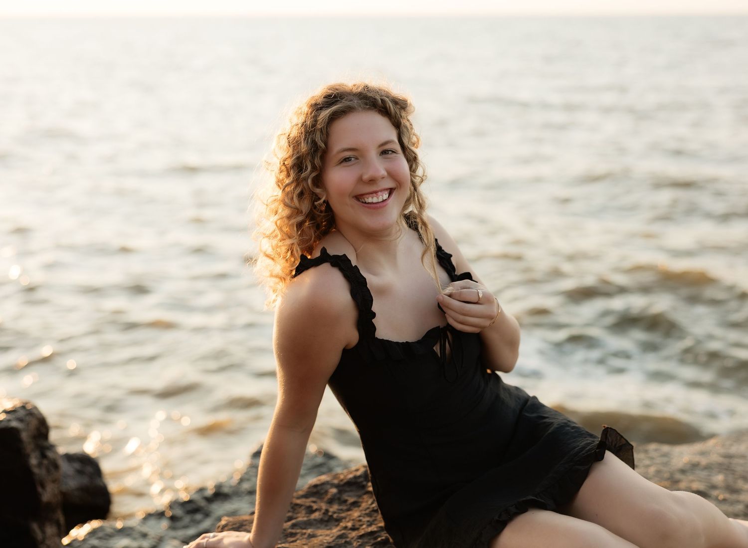 Smiling woman with curly blonde hair sitting on rocks by the ocean at sunset in a black dress.