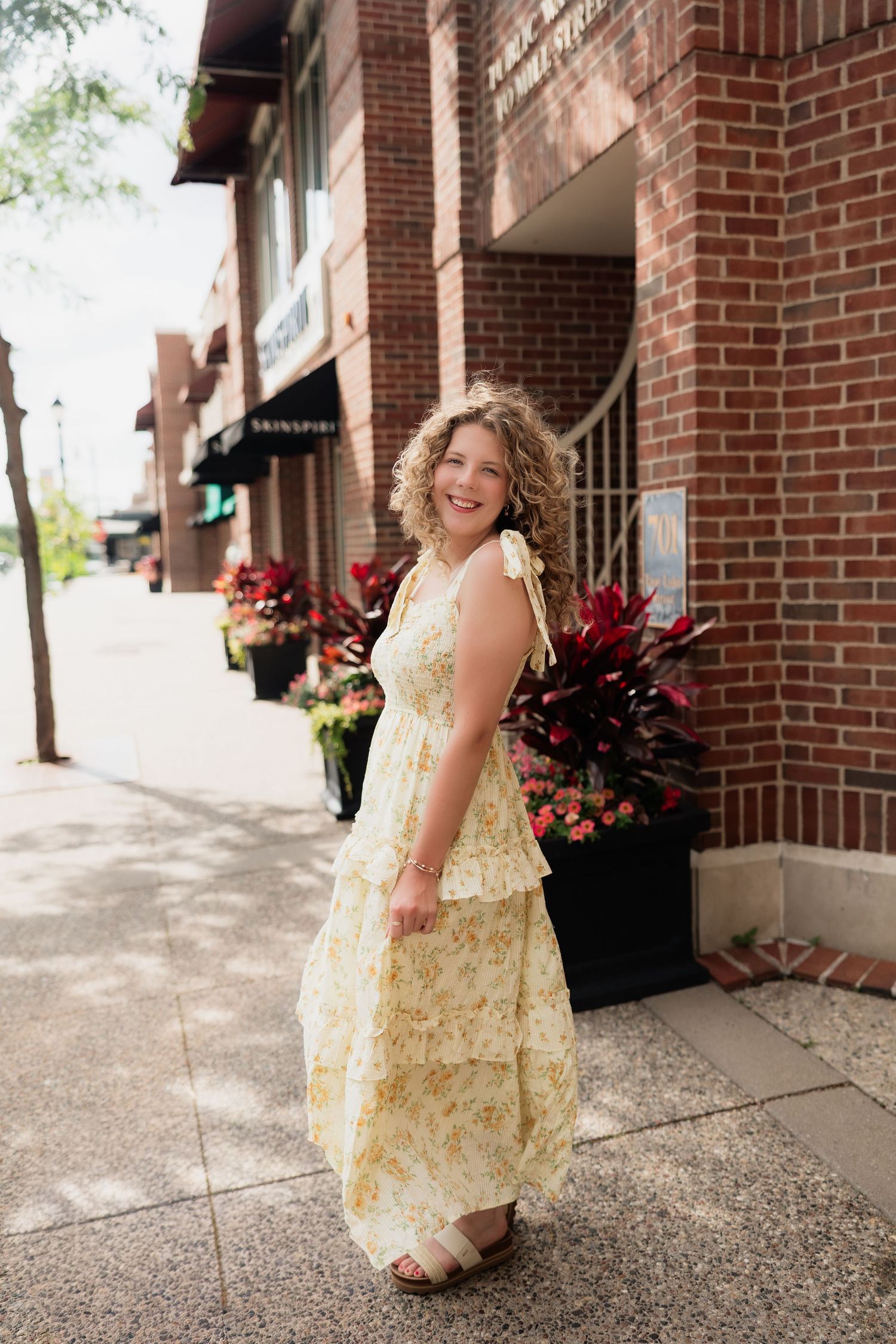 Young woman in yellow floral tiered maxi dress smiling on urban sidewalk near brick building with flowers.