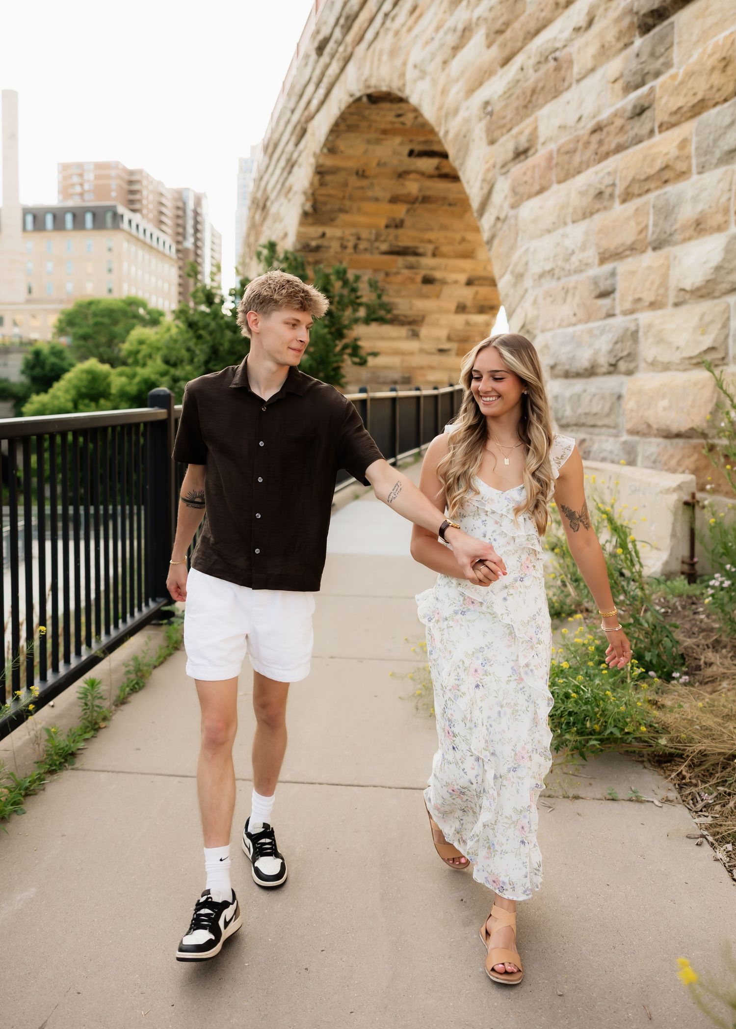 Couple walking hand in hand along a stone arch bridge pathway in Minneapolis, woman in floral dress, man in black shirt.