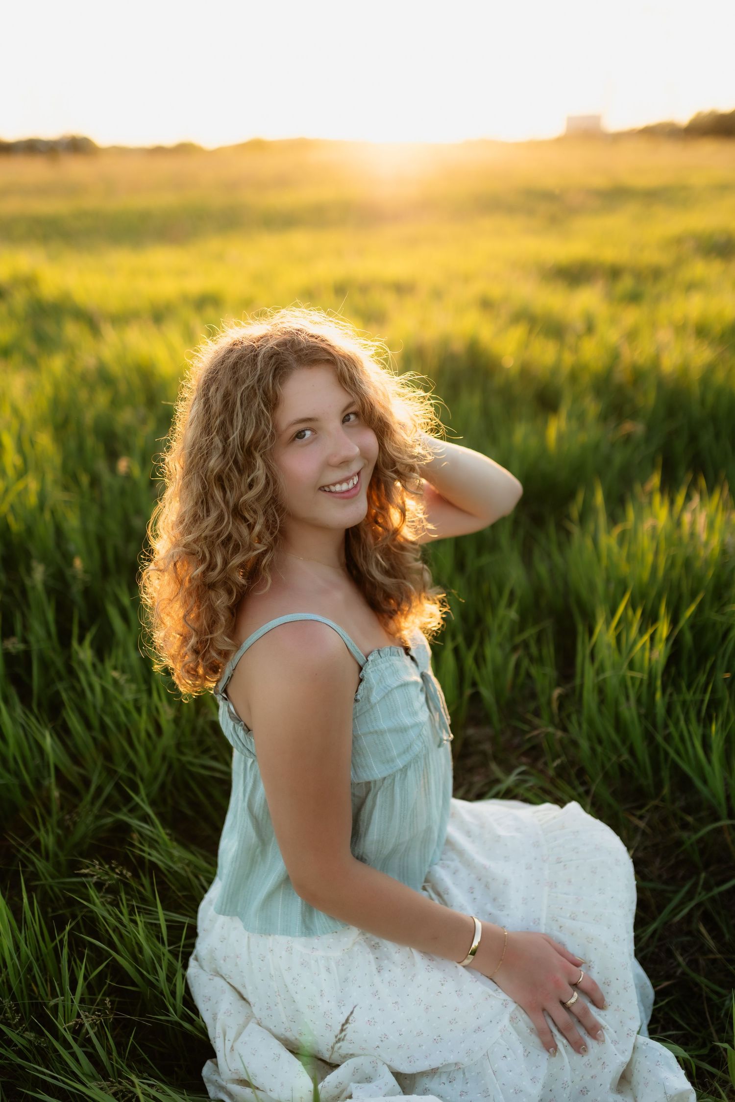 Young woman with curly hair in a light blue dress sitting in a green field during golden sunset.
