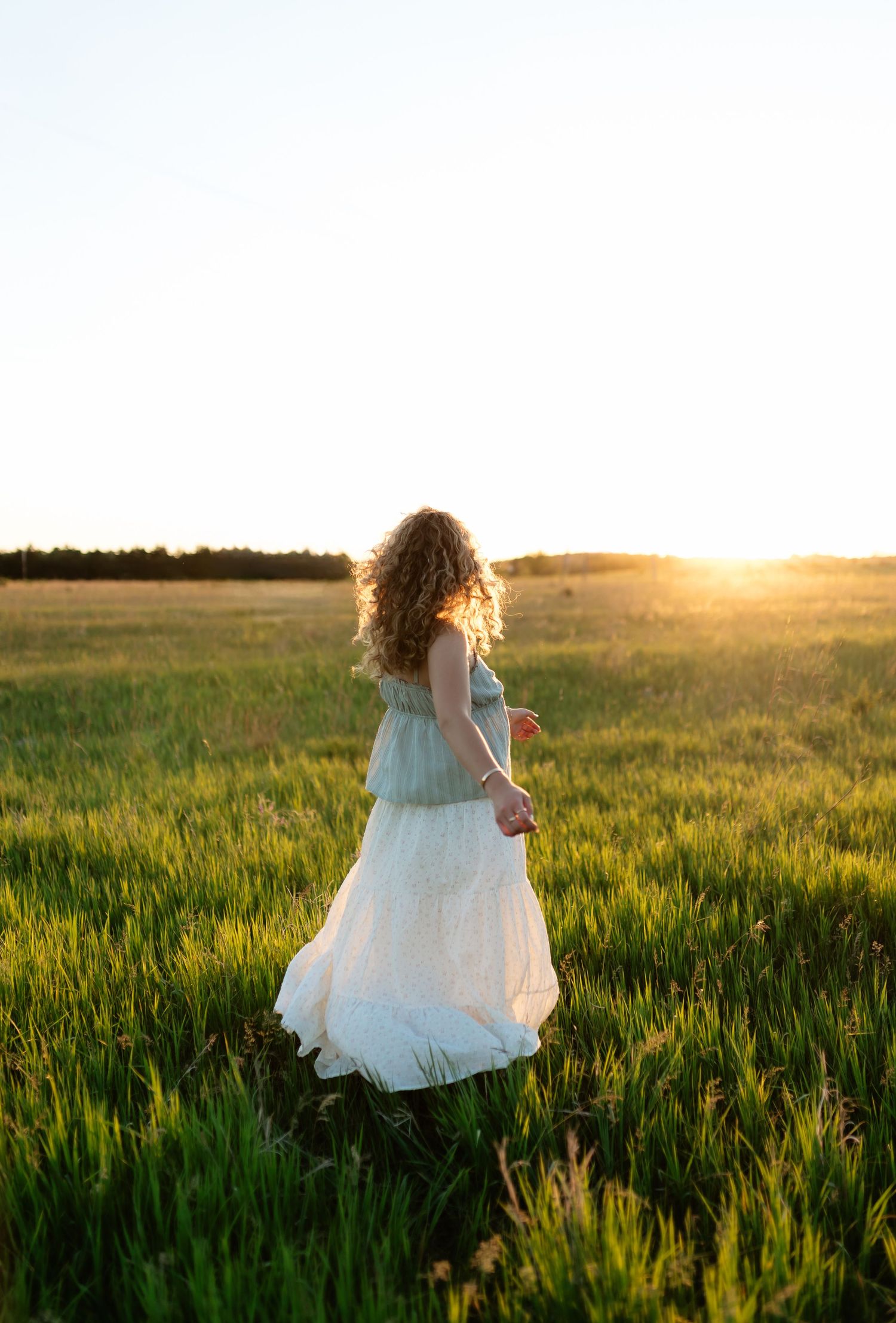 A woman in a white skirt and blue top walks through a green field at golden sunset with curly hair flowing.