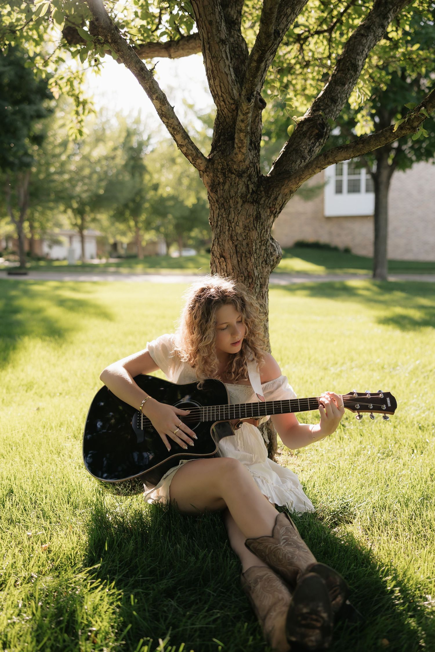 Young woman with curly hair playing acoustic guitar while sitting against a tree on green grass outdoors.