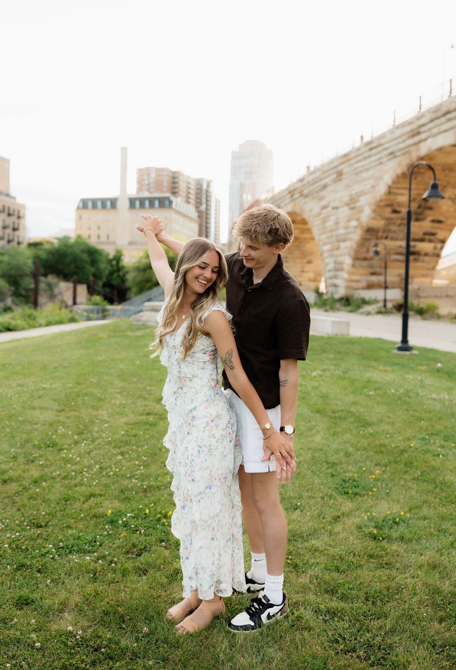 Happy couple laughing on green lawn with historic stone arch bridge and city skyline behind them.