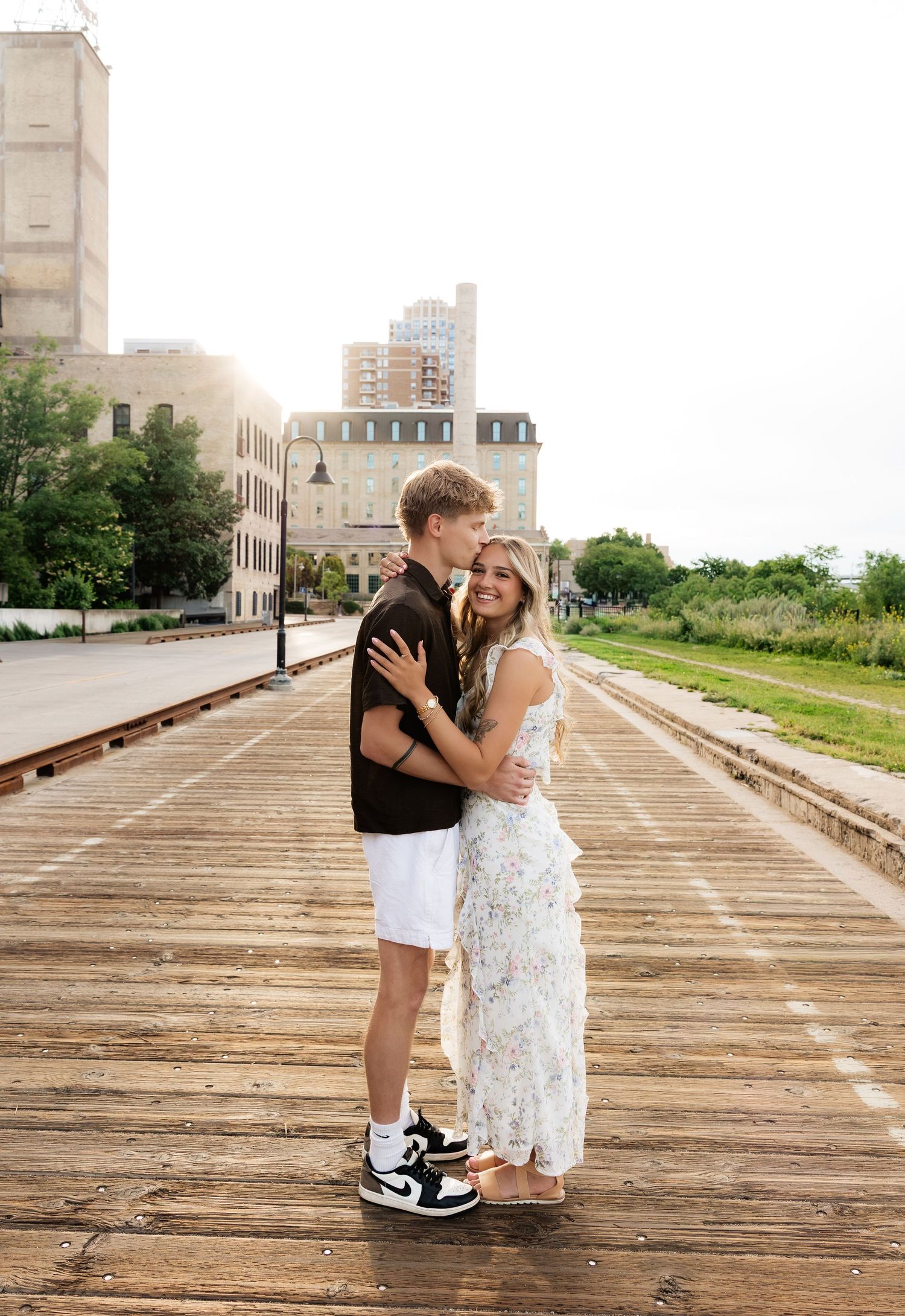 Couple embracing and kissing on a wooden boardwalk with urban buildings in the background.