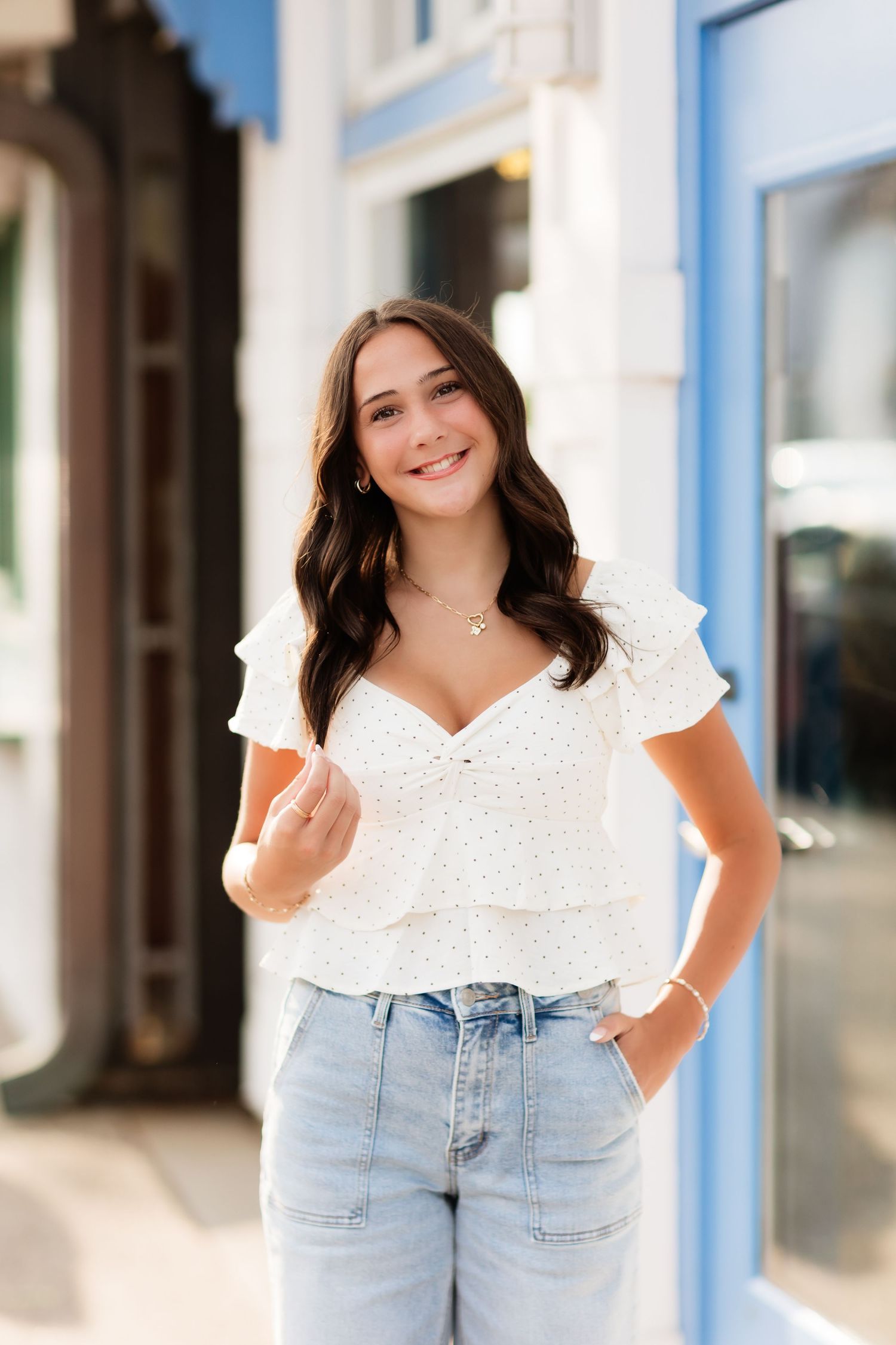 Young woman in white ruffled top and light wash jeans smiling outdoors near blue storefront.