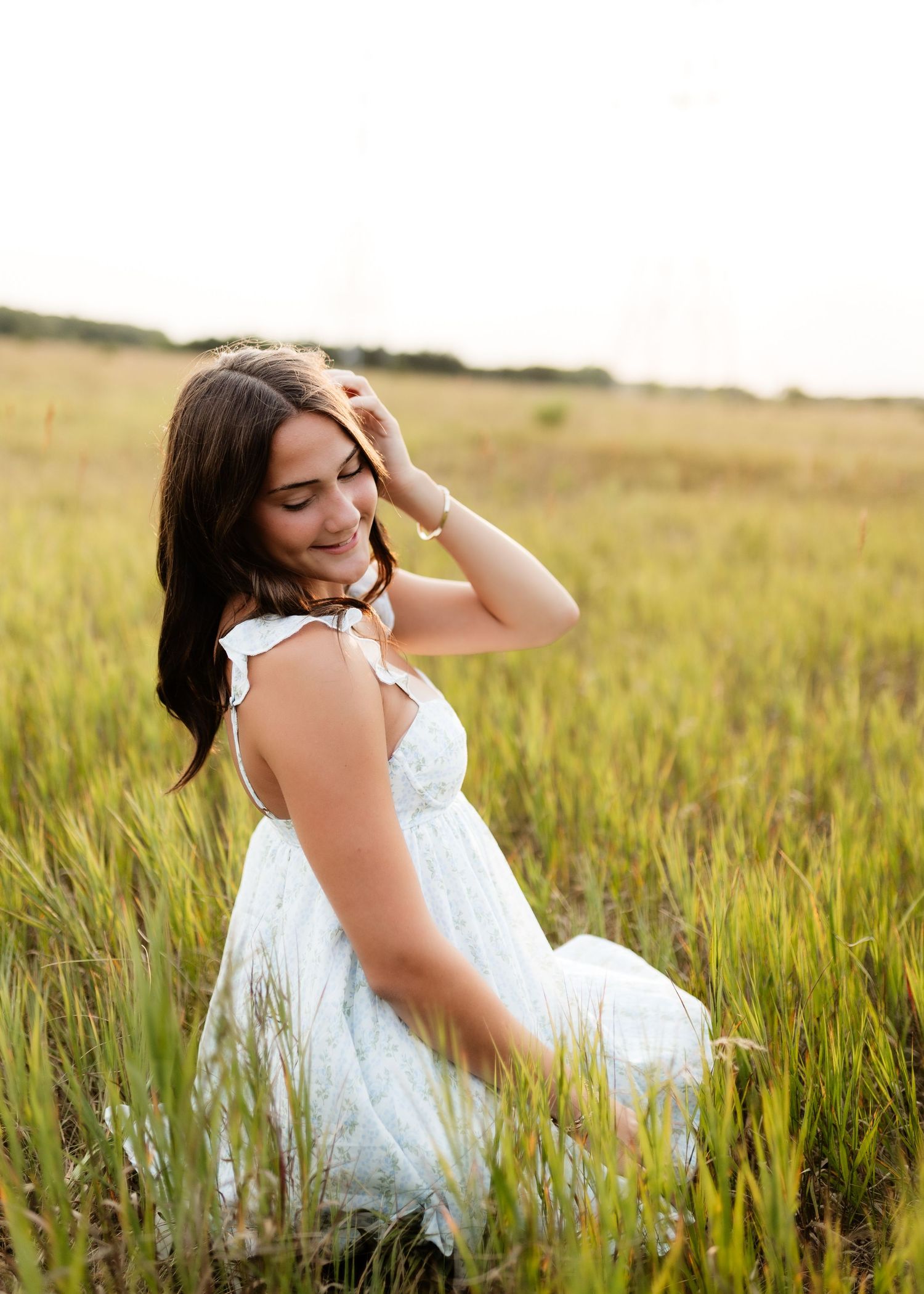 Young woman in white dress sitting in tall grass field at golden hour.