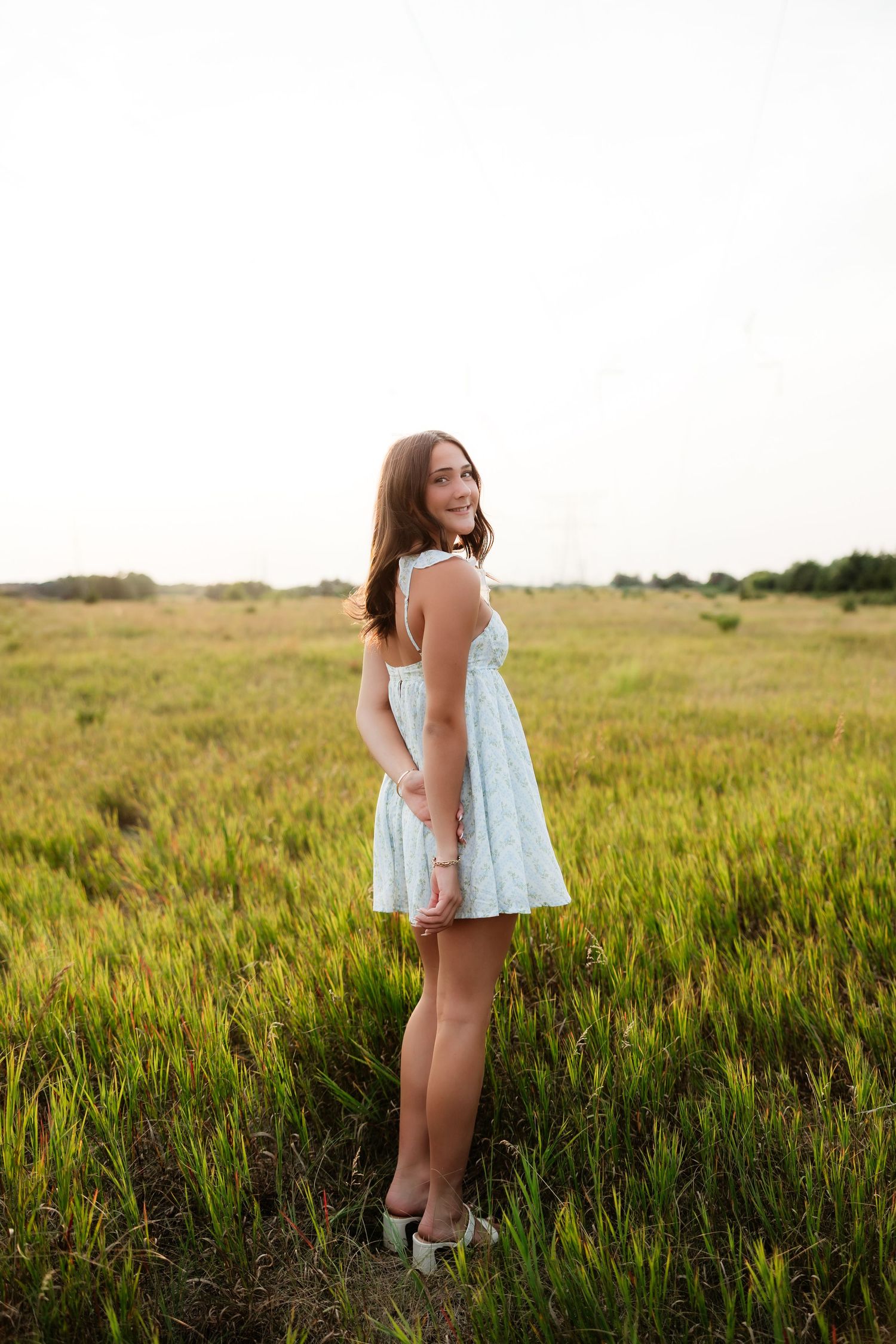 Young woman in light blue mini dress standing in open green field at dusk.
