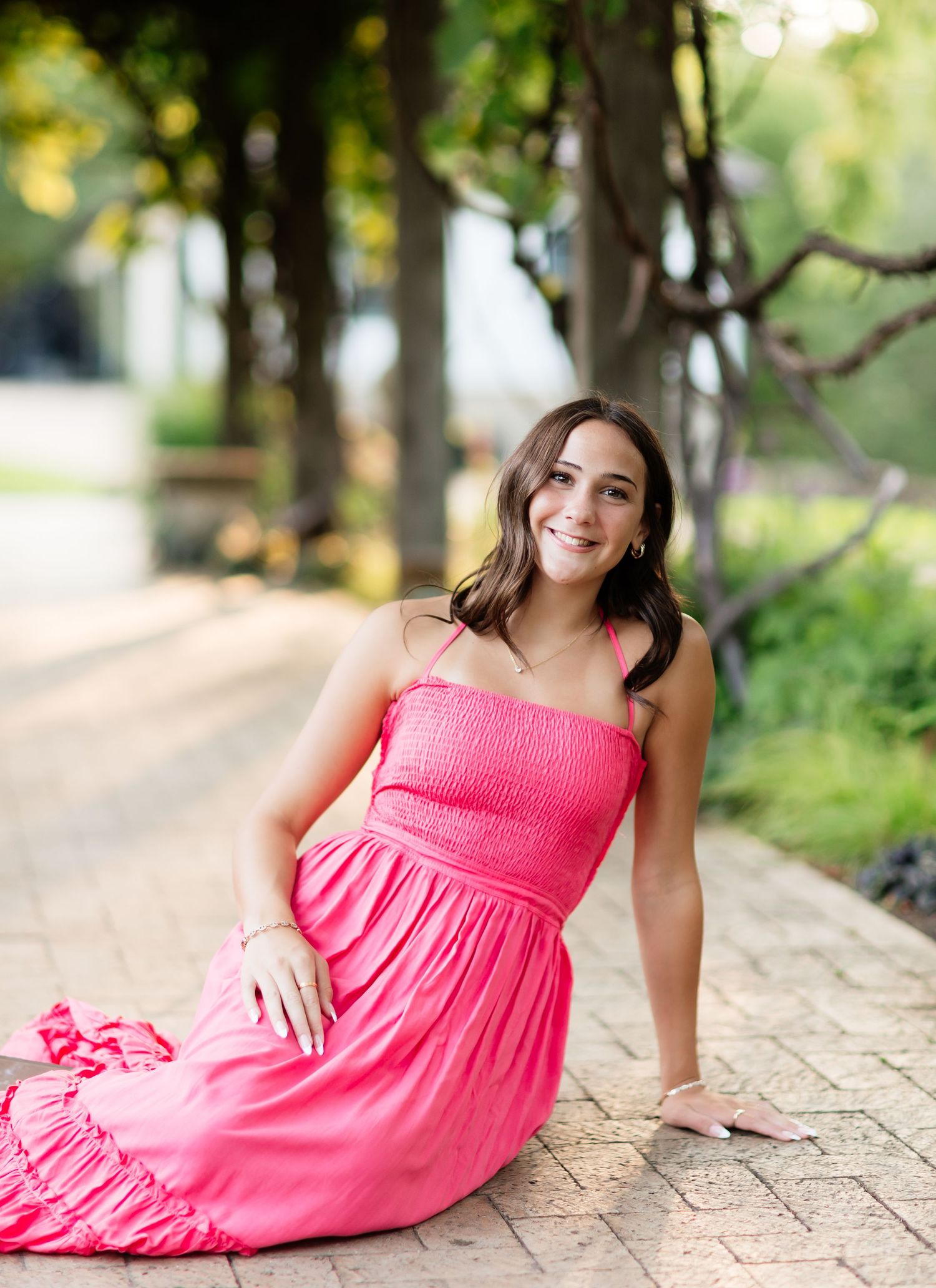 Smiling young woman in hot pink dress sitting on brick pathway in park.