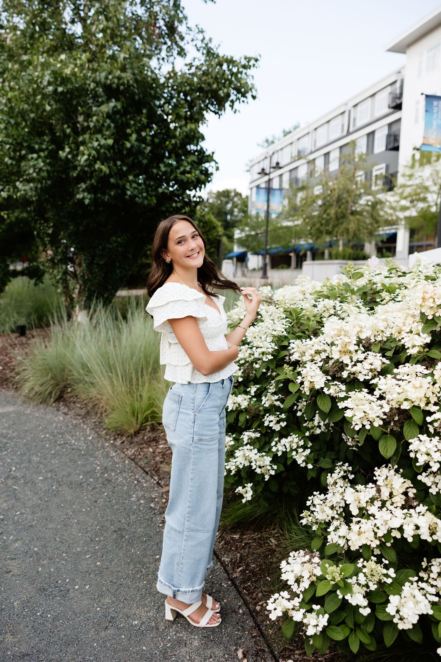 Smiling brunette woman in white ruffled top touching white hydrangea blooms in outdoor urban garden.