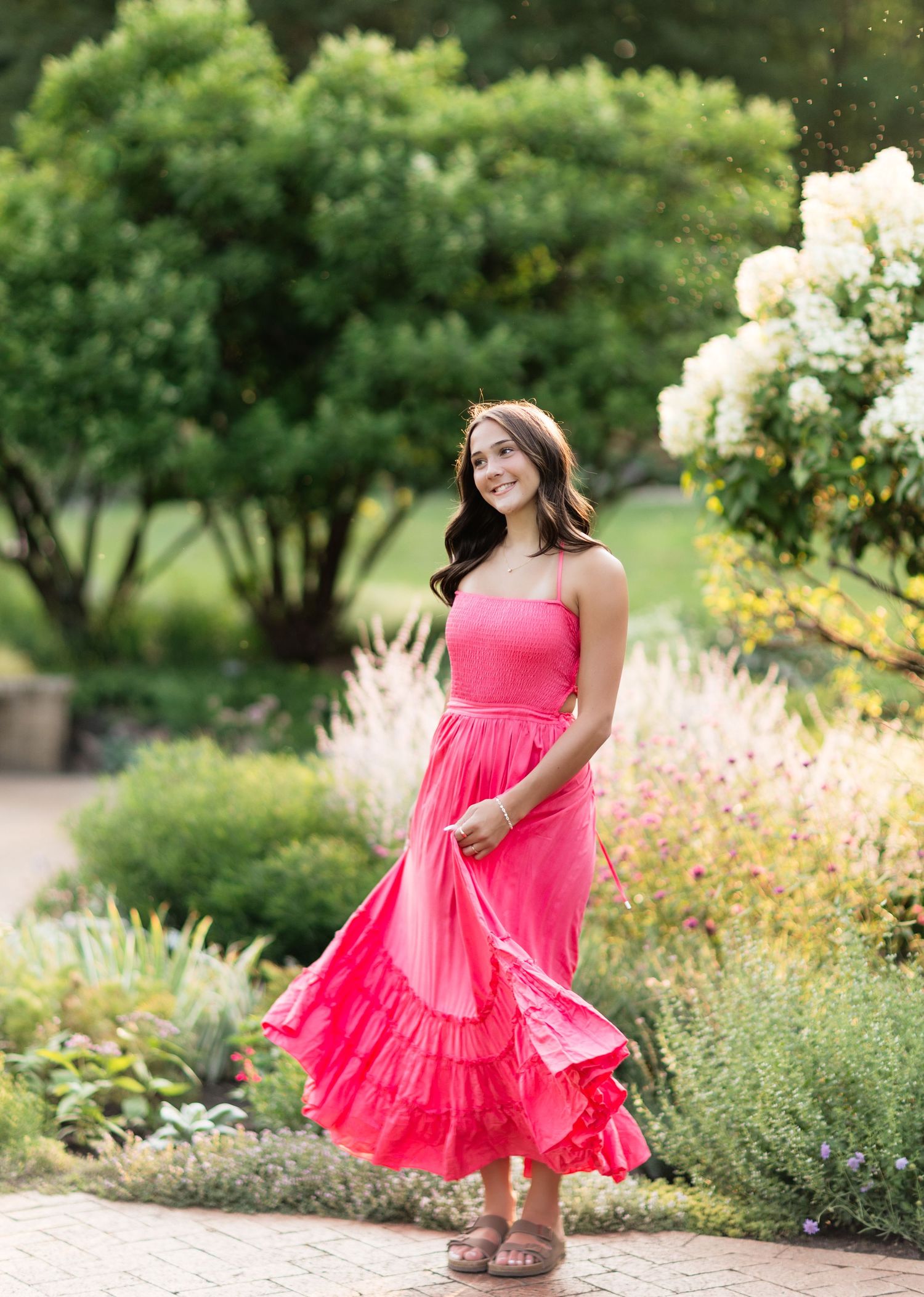 Young woman in pink maxi dress twirling in a lush garden with colorful flowers and green trees.