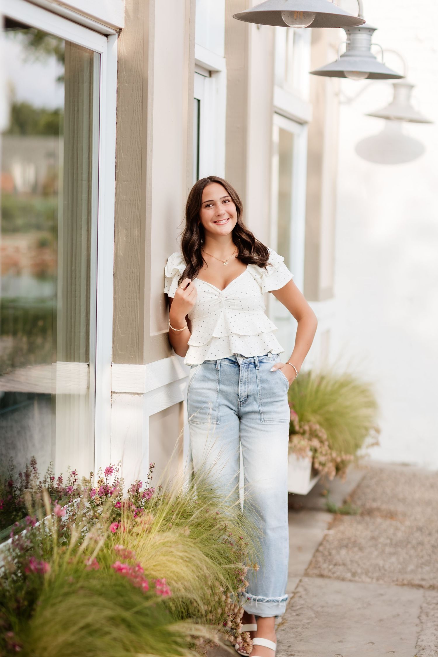 Smiling young woman in white ruffle top and distressed jeans leaning against a building with pink flowers nearby.