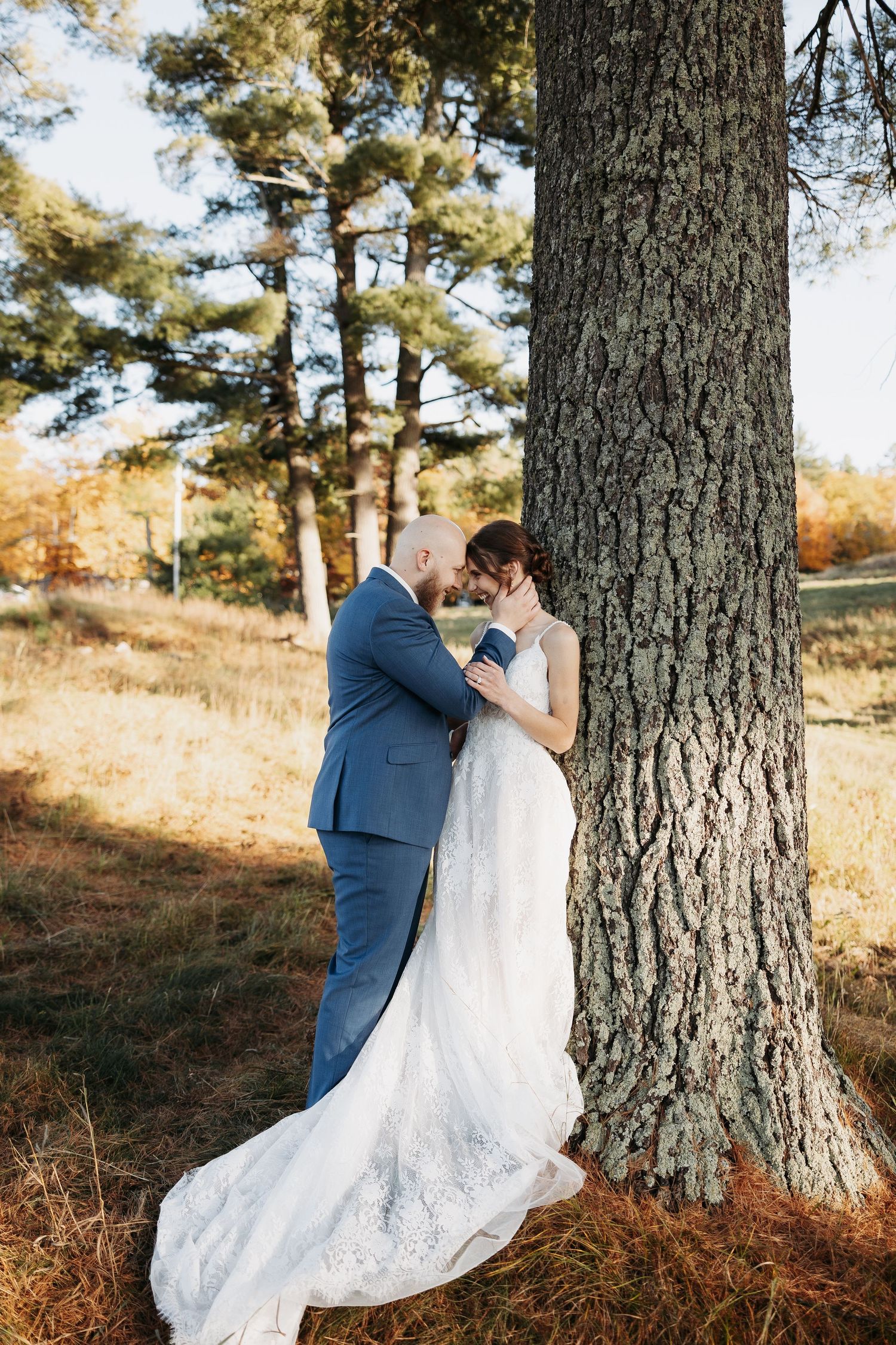 Bride and groom sharing an intimate moment leaning against a large tree in an autumn forest setting.