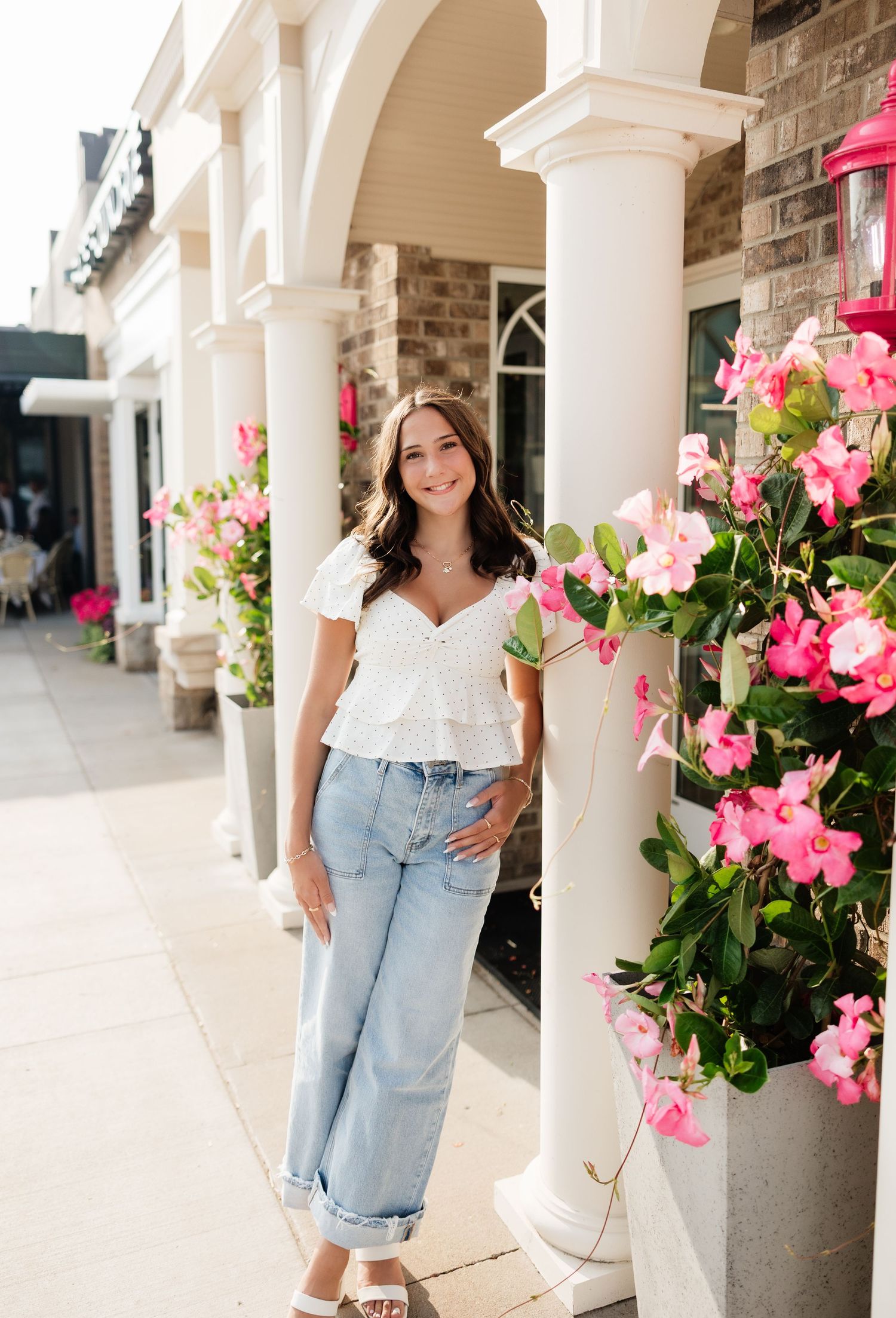 Smiling young woman in white top and rolled jeans poses among vibrant pink flowers by white columns.