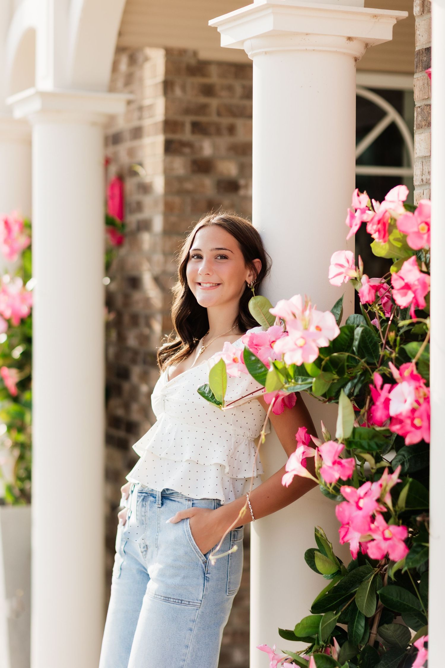 Young woman in white top and jeans smiling among pink flowers by white columns.