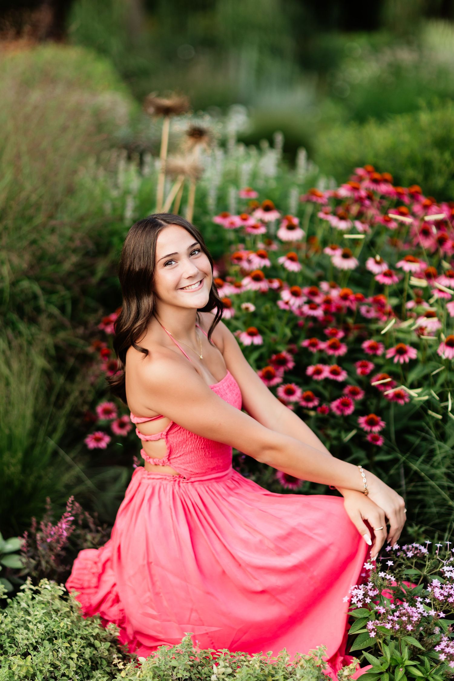 Smiling young woman in pink dress sitting among blooming echinacea flowers in a garden setting.