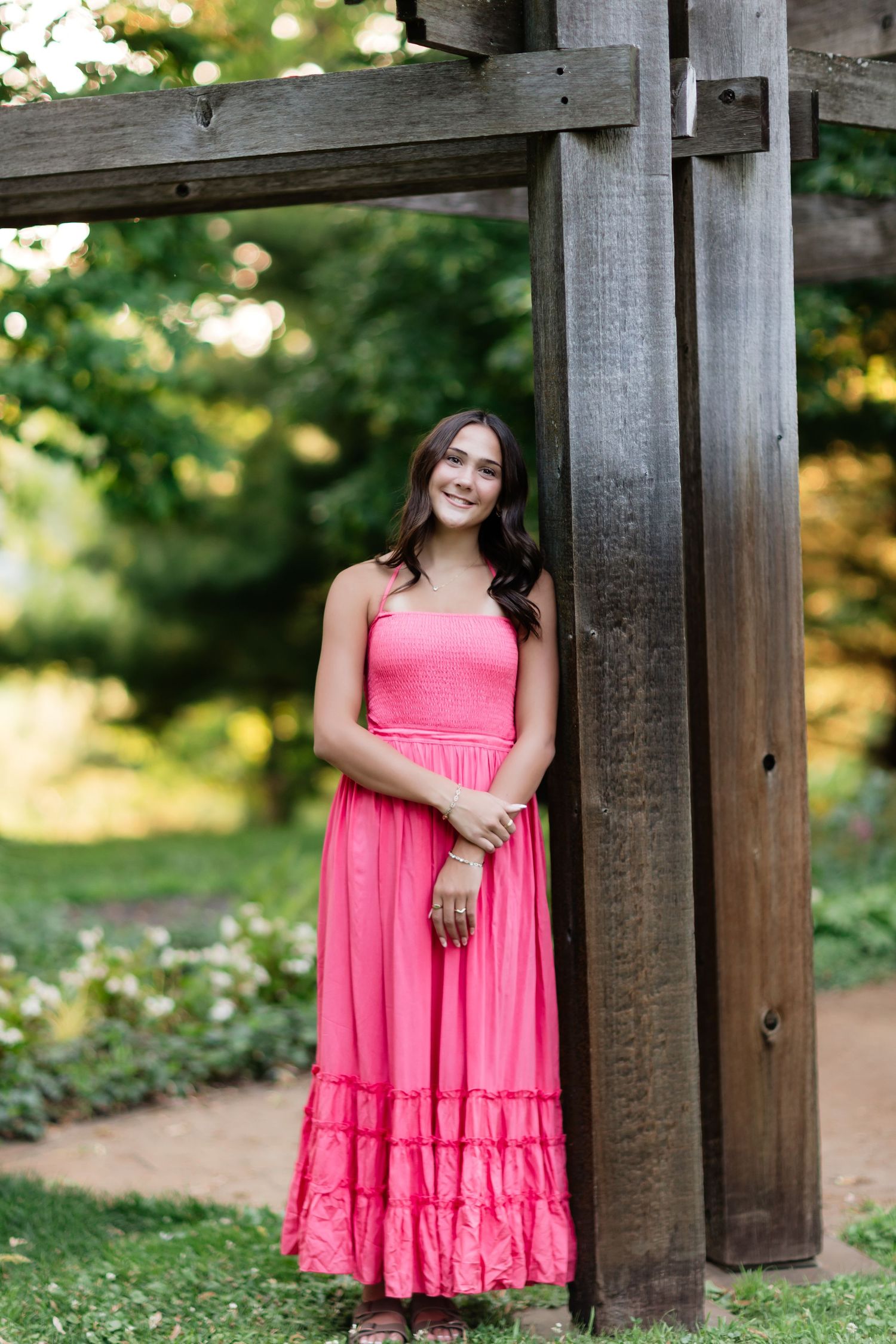 Smiling brunette teen in pink maxi dress leaning against a wooden pergola post in a lush garden.