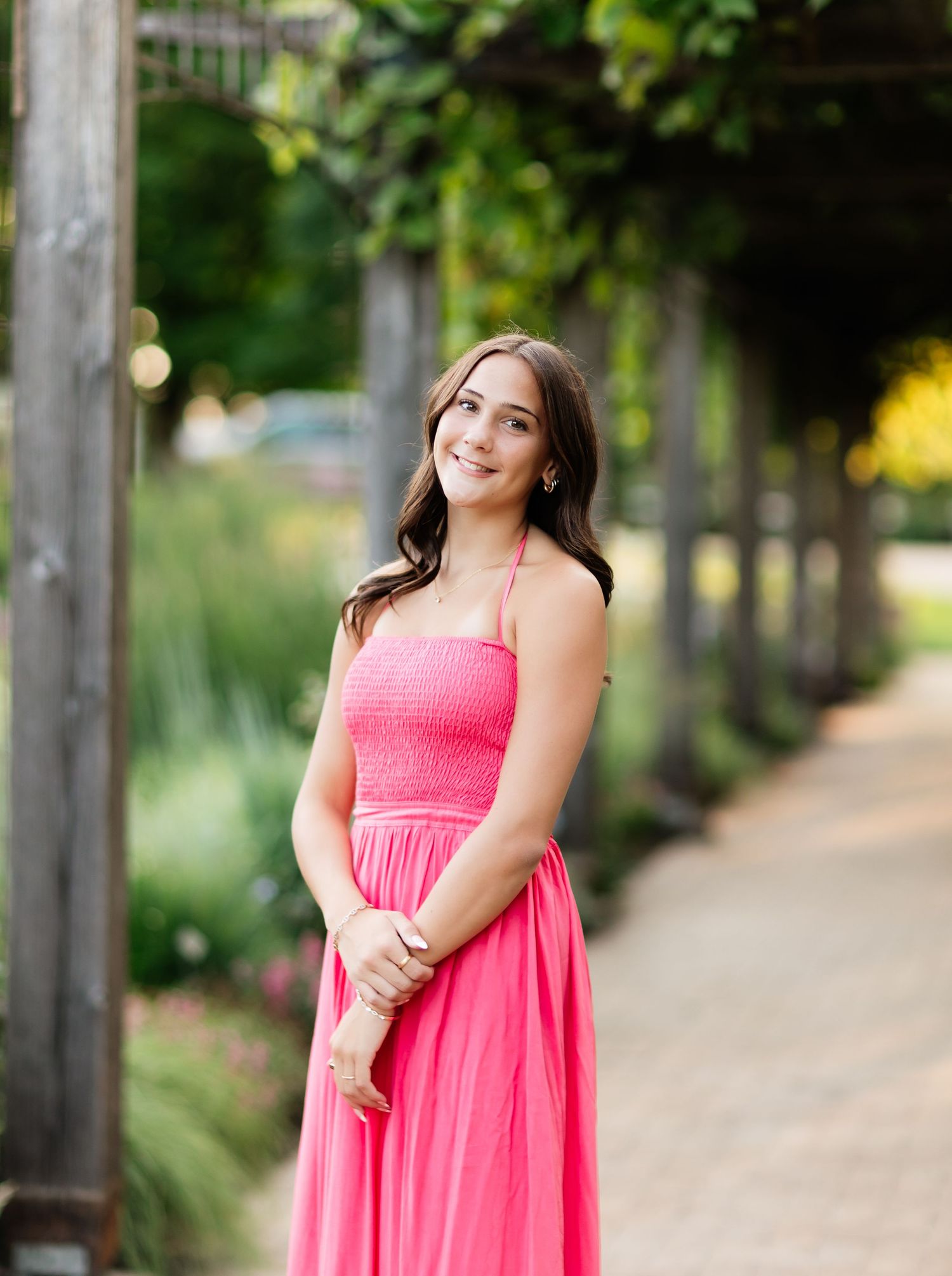 Young woman in bright pink smocked maxi dress smiling under green garden pergola pathway.