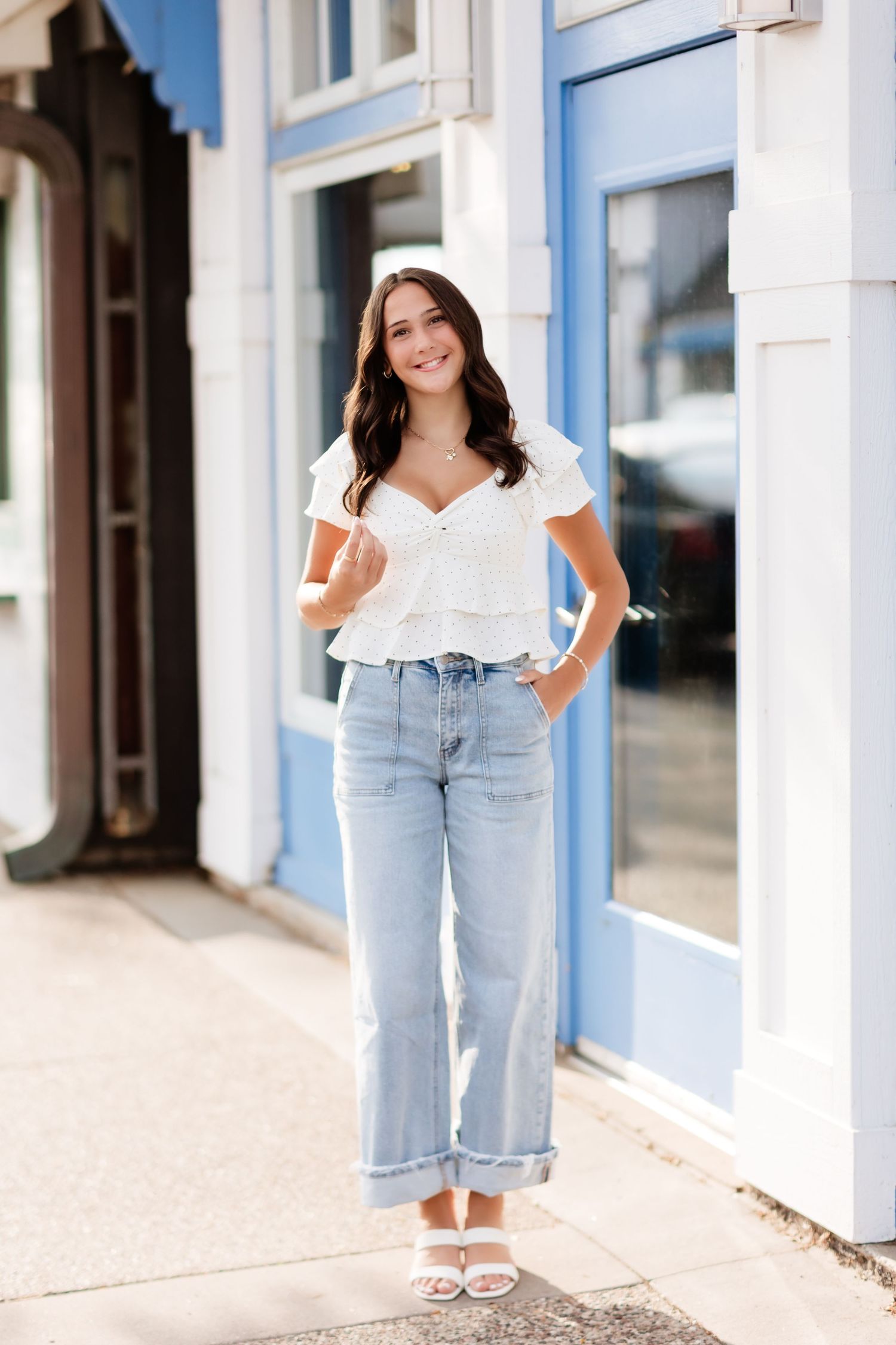 Smiling brunette woman in white ruffle crop top and light wash wide-leg jeans stands outside blue storefront.