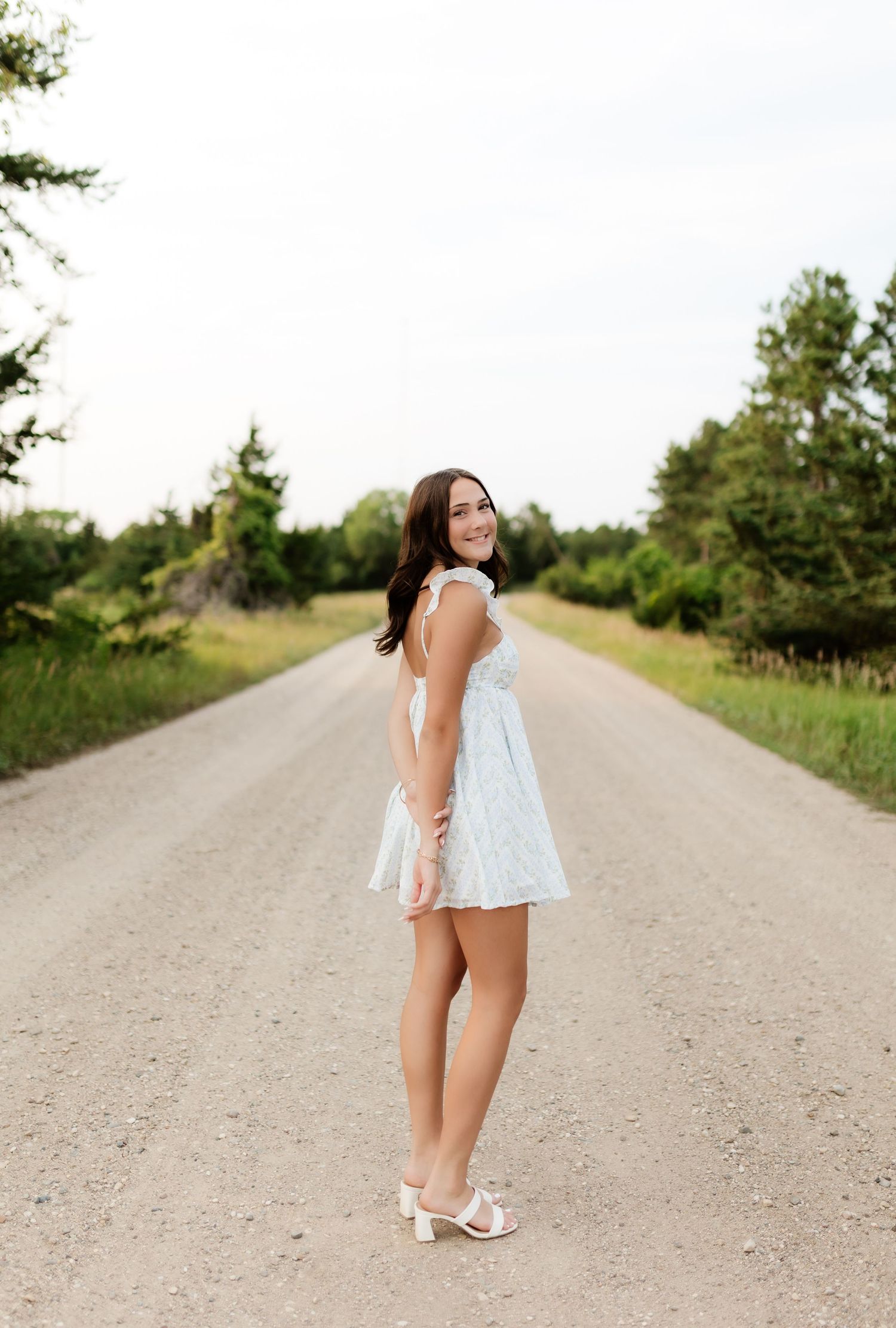 Young woman in white mini dress and heels smiling on a rural gravel road surrounded by green trees.