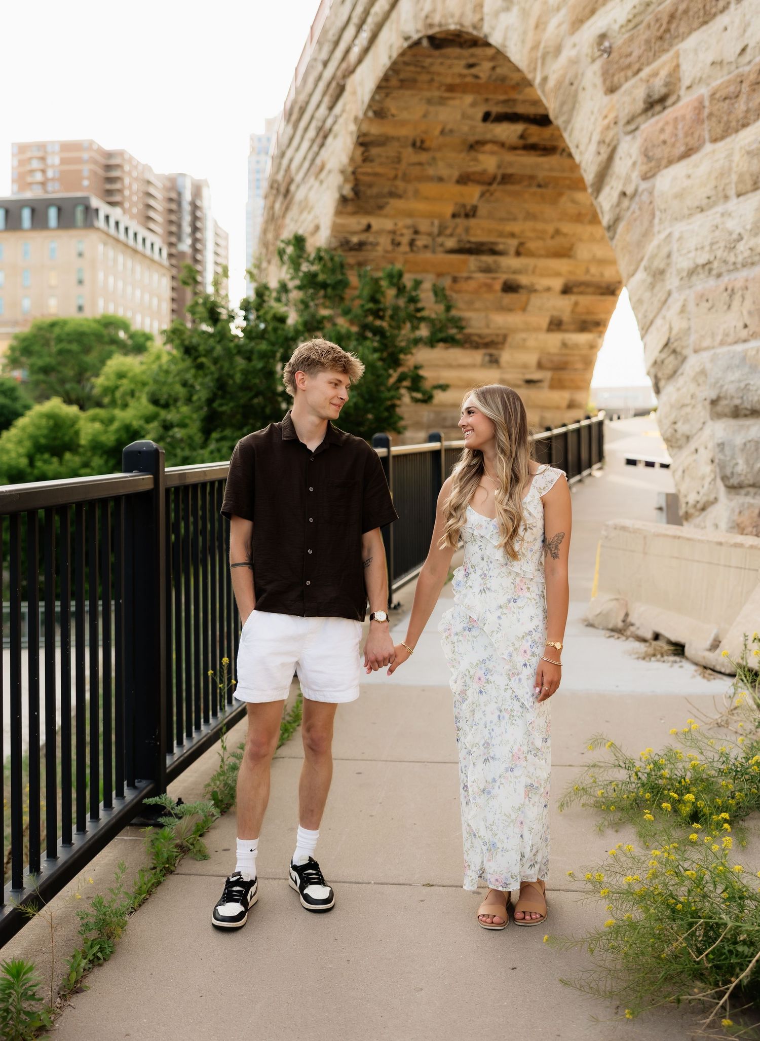 Young couple holding hands and walking along a stone arch bridge pathway in an urban setting.
