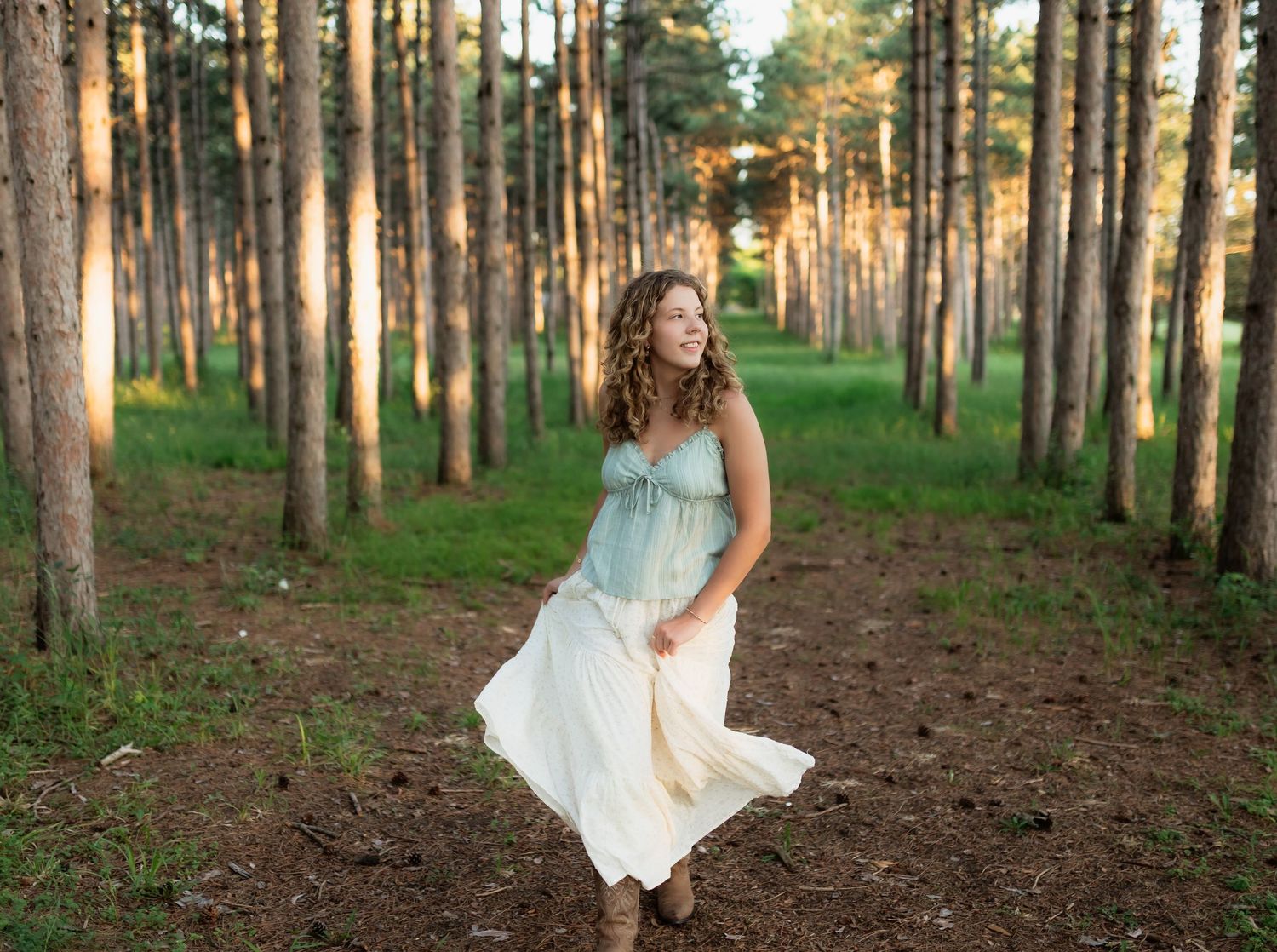 Young woman in flowing white and blue dress walking through a pine forest in golden evening light.