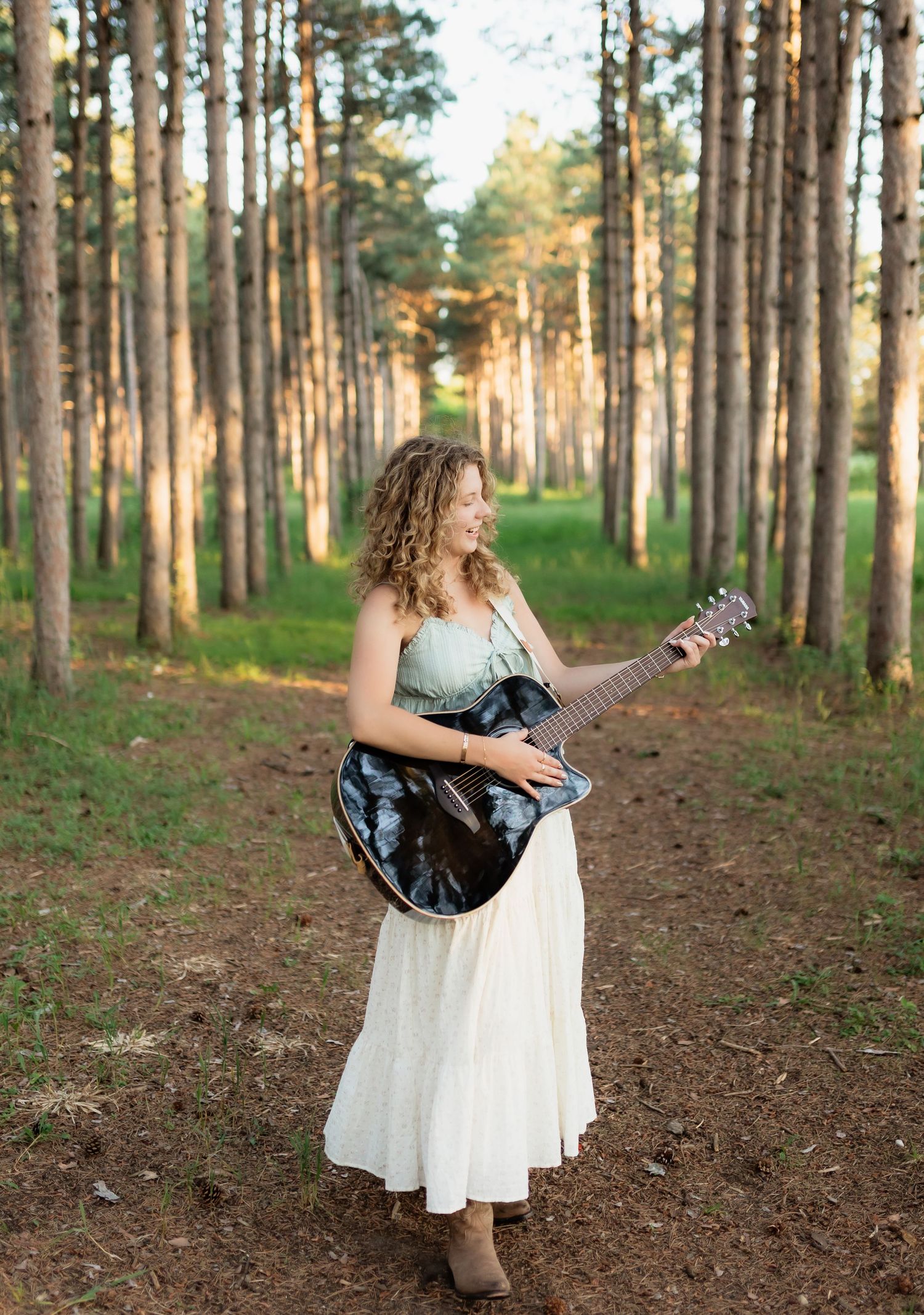Woman with curly hair playing guitar on a forest path in a white dress at golden hour.