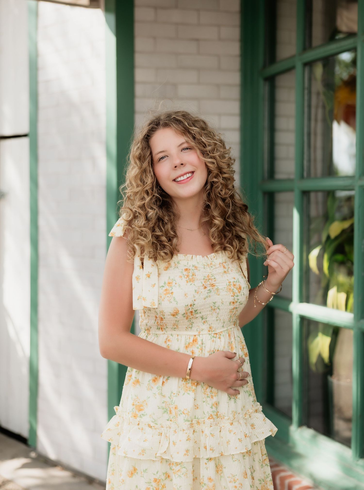Smiling teenage girl with curly hair wearing a yellow floral sundress near a green window.