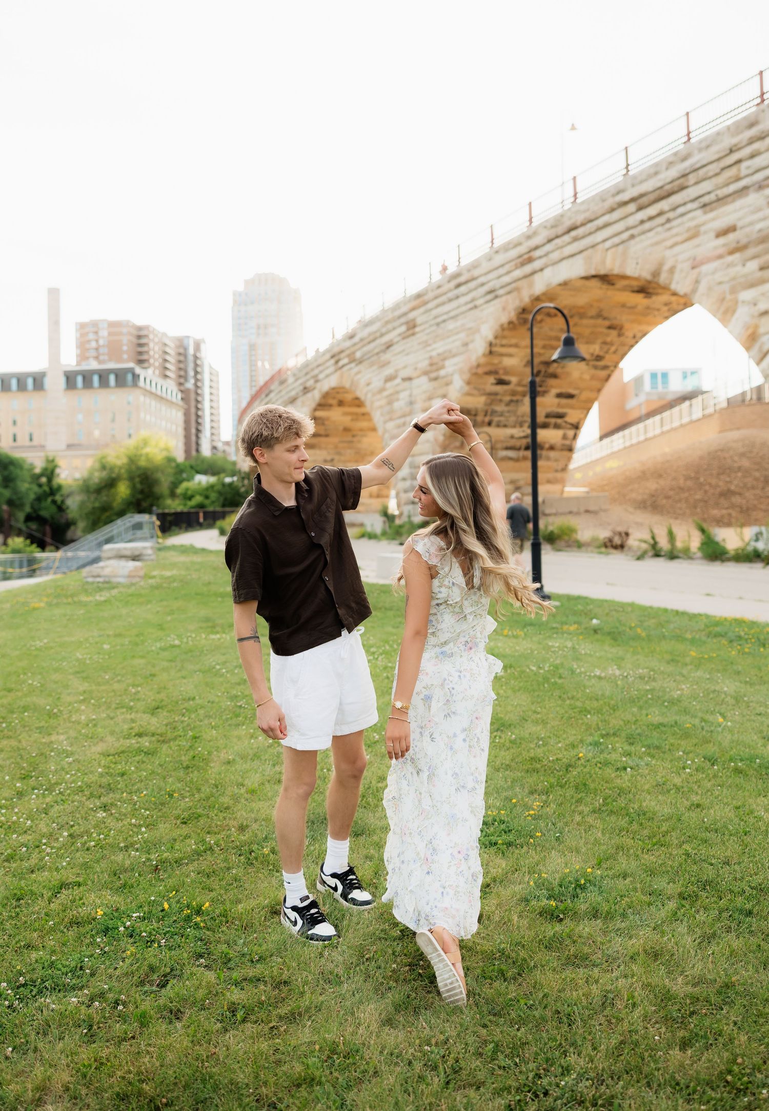 A couple dances near Stone Arch Bridge in Minneapolis, MN, with the young man spinning the woman in a floral dress.