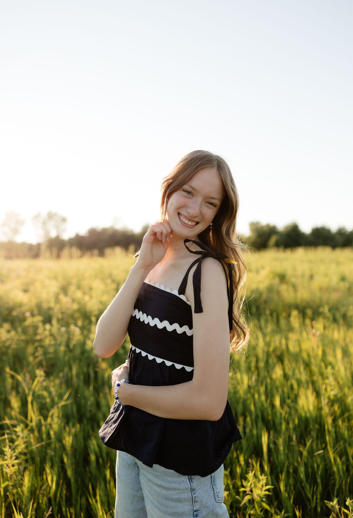 Smiling young woman in black top with white trim standing in a sunlit green field.