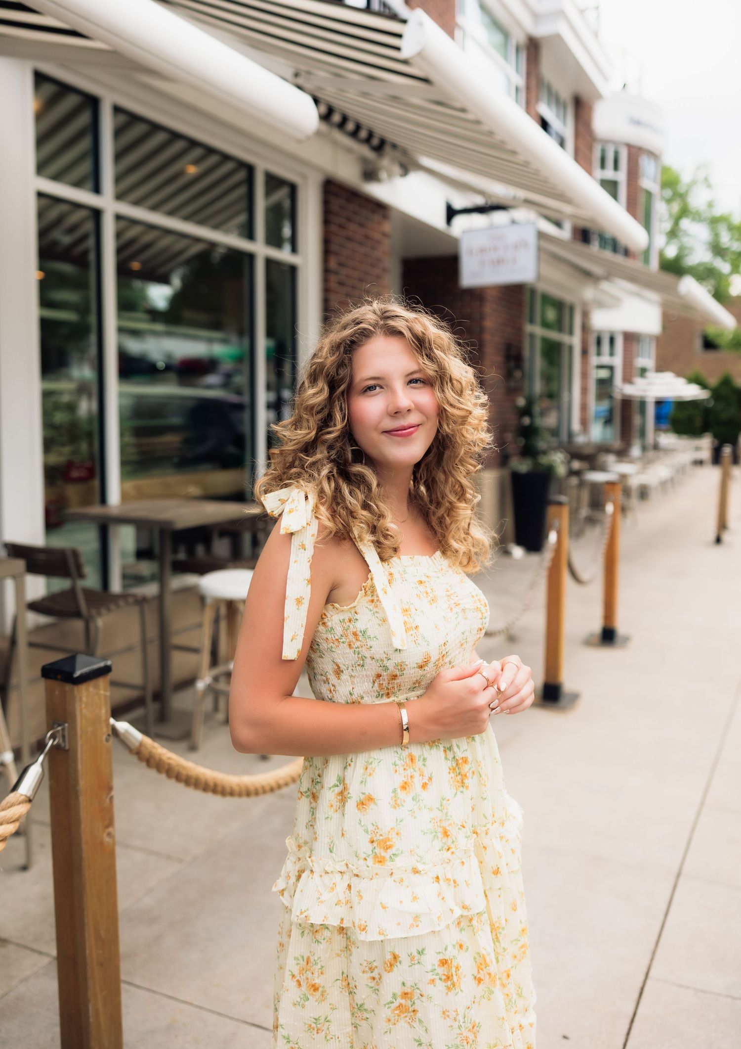 Young woman with curly hair in floral sundress standing on urban sidewalk outside restaurant.