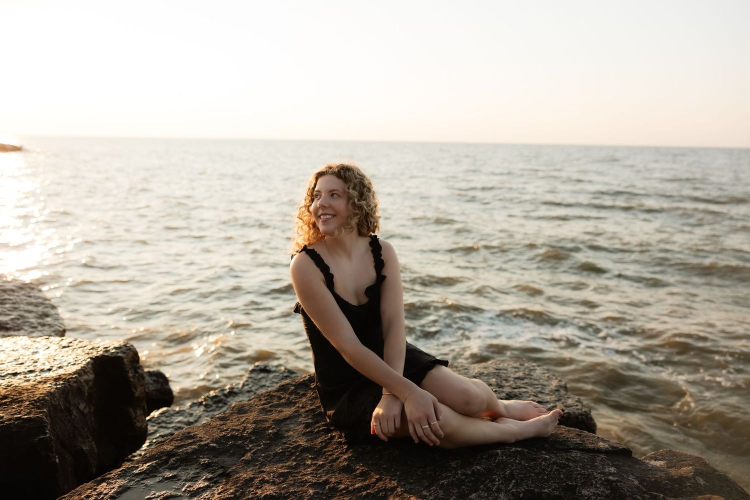 A smiling woman in a black dress sits on rocky shore by the sea at sunset, enjoying a peaceful moment.