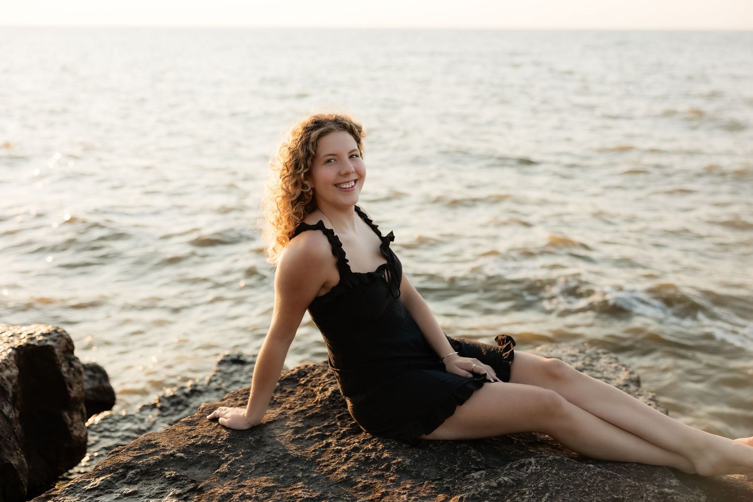 Smiling woman with curly hair in black dress sitting on rocks by the ocean at sunset.