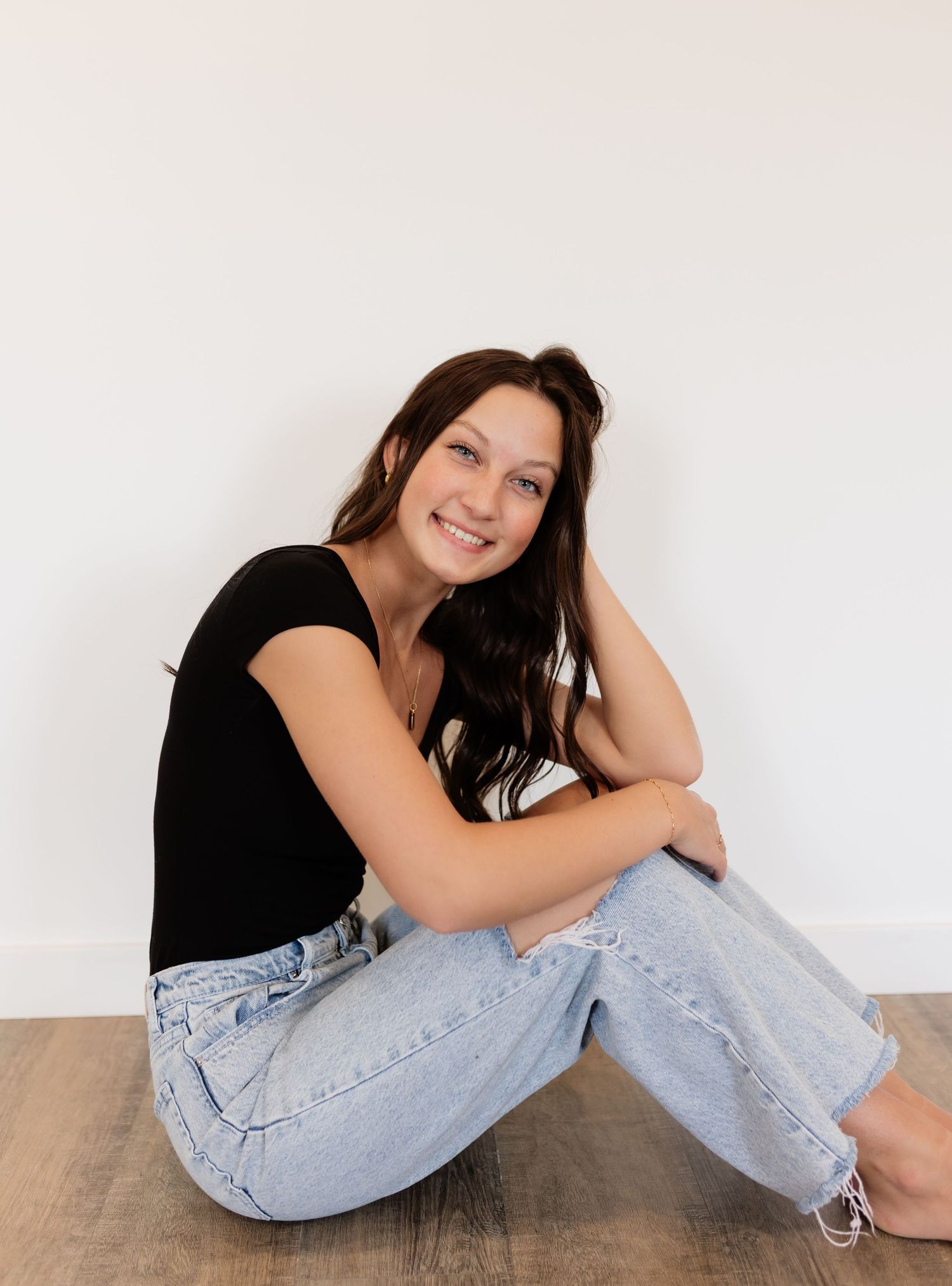 Young woman with dark hair sitting on floor, smiling, wearing black top and light blue jeans against white wall.