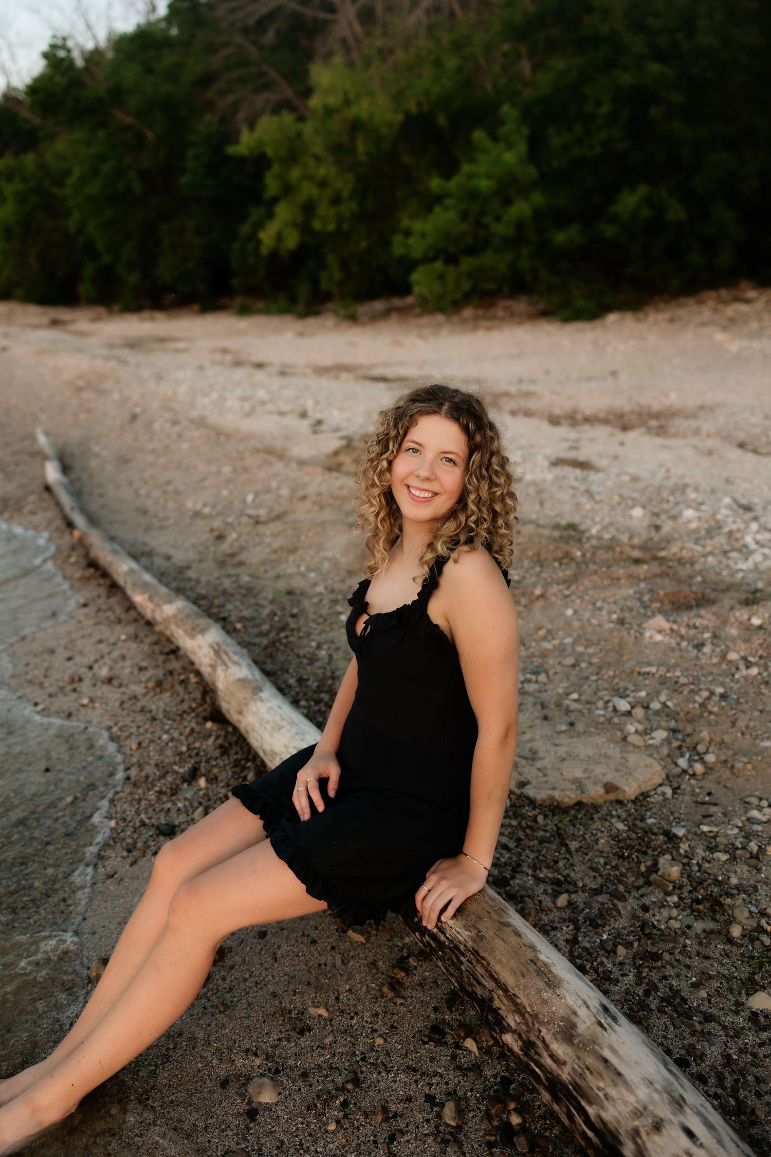 Smiling woman in black dress sitting against driftwood log on a sandy beach at golden hour.