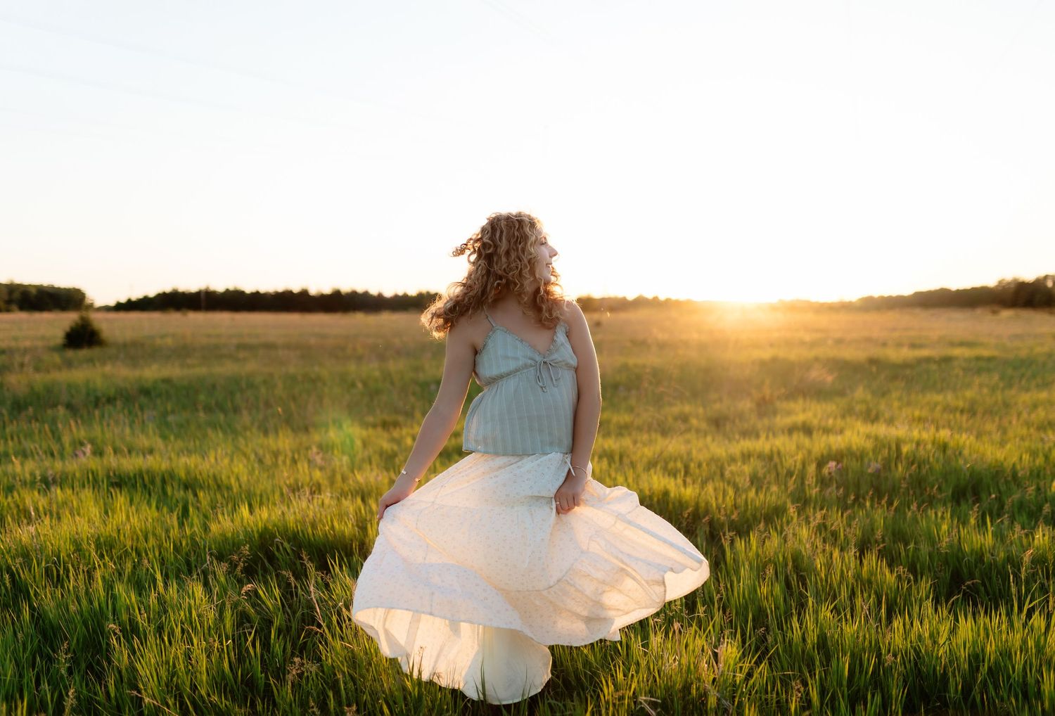 Young woman in a light blue top and white skirt twirling in a sunlit green field at sunset.