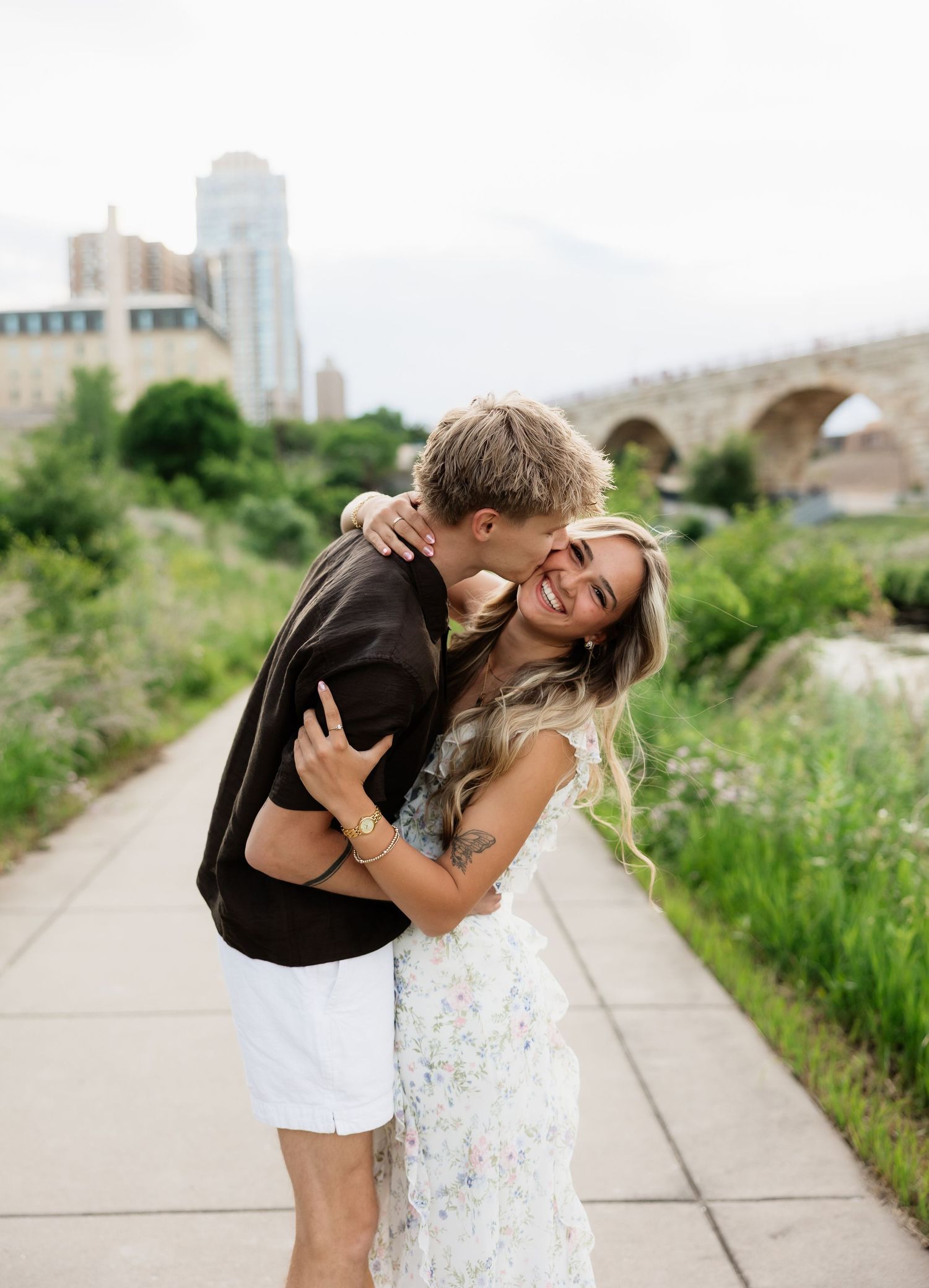 Joyful couple embracing on pathway with lush greenery and stone arch bridge in background.