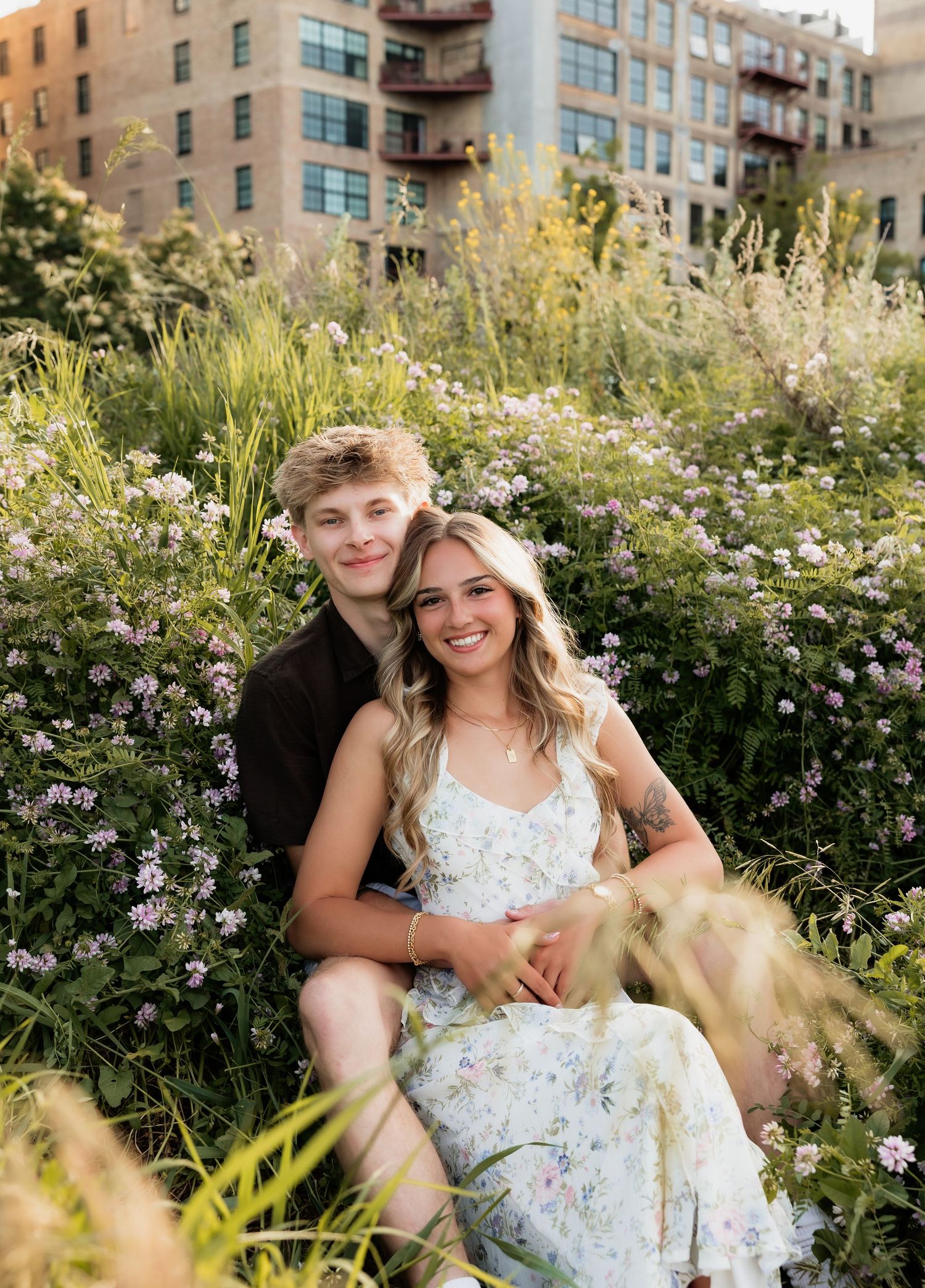 Couple sitting together in a wildflower meadow with urban buildings in the background on a sunny day.