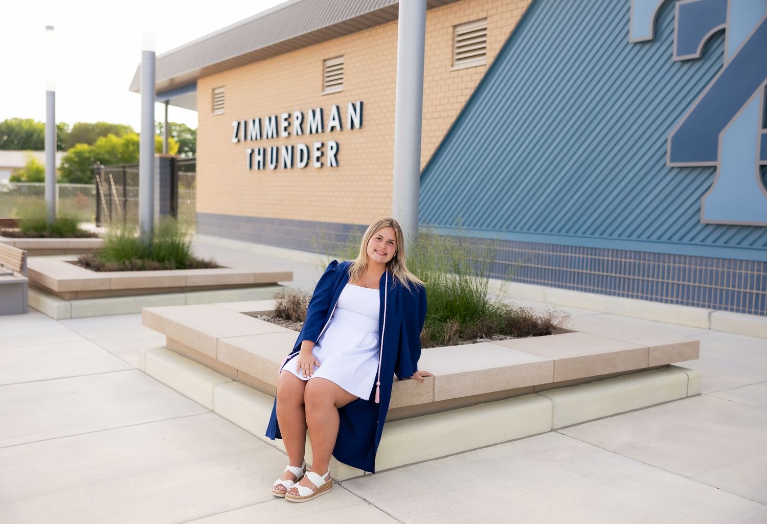 Smiling graduate in a blue gown posing in front of Zimmerman Thunder high school.