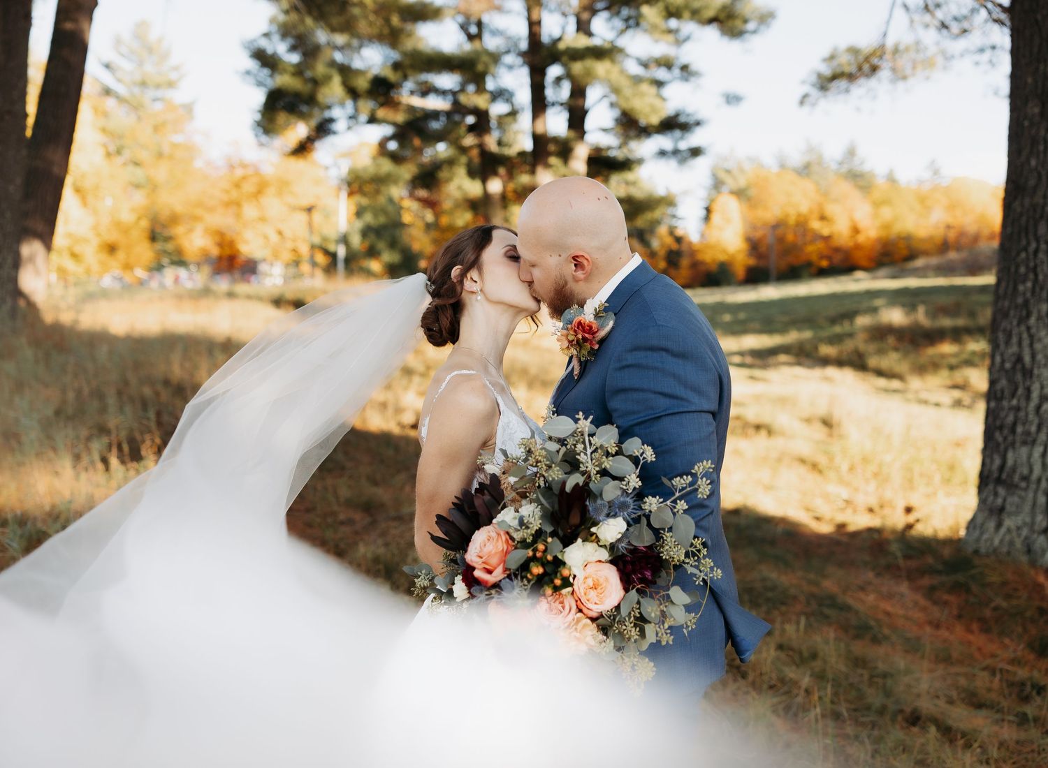 Bride and groom kissing under a flowing veil with autumn trees and colorful fall foliage in background.