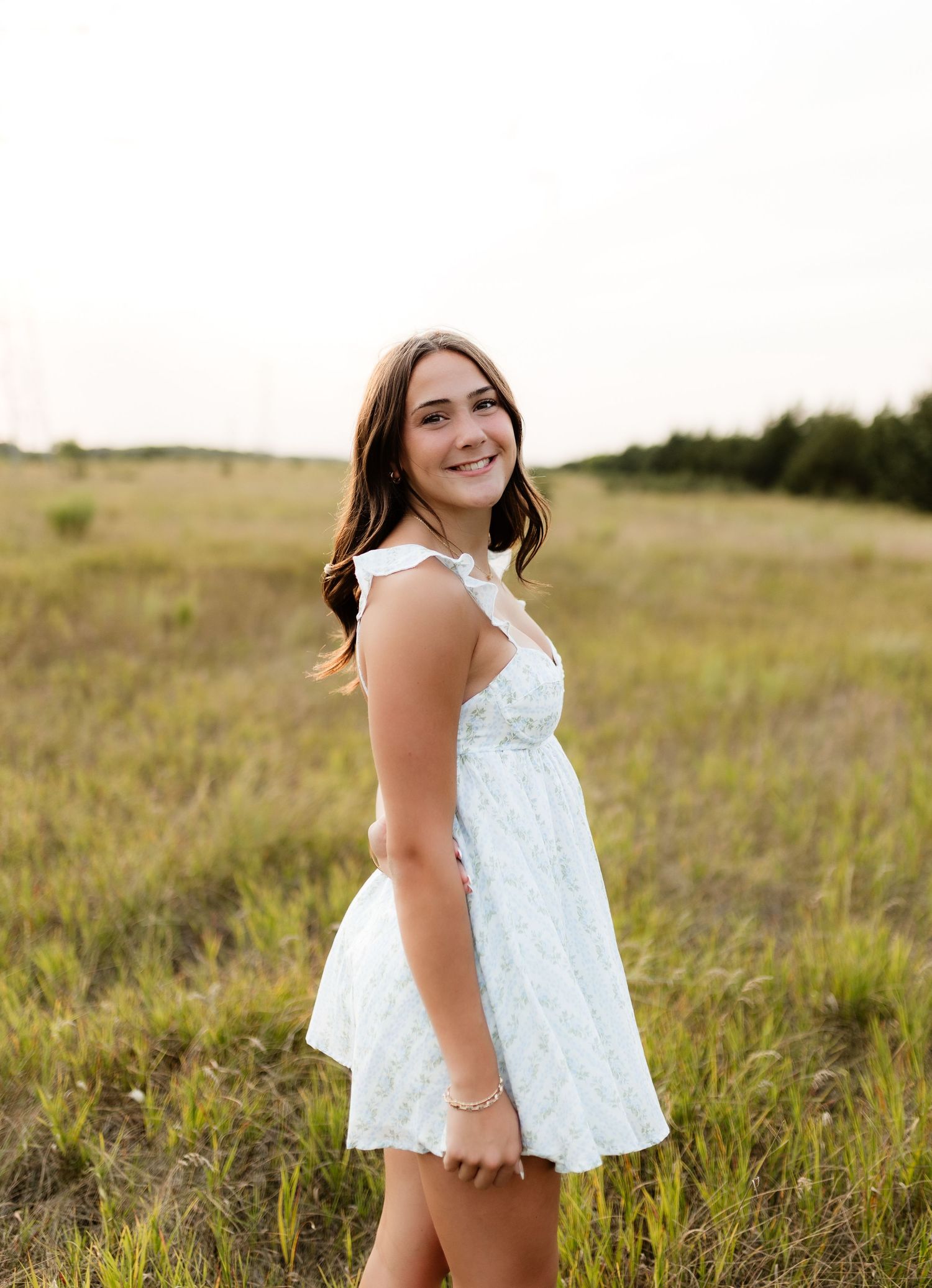 Smiling young woman in a light blue floral mini dress standing in an open grassy field.